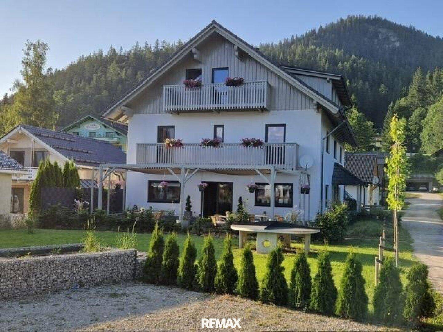 Traditionelles Haus mit mehreren Balkonen, einer Terrasse und gepflegtem Garten vor einer Bergkulisse.