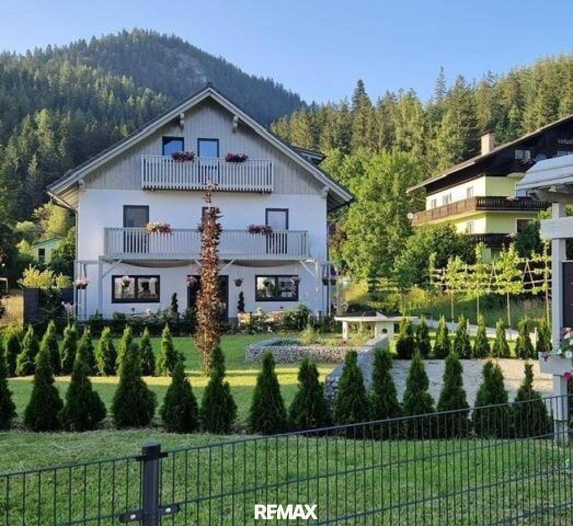 Gepflegtes Einfamilienhaus mit Balkonen und einer Gartenanlage, umgeben von grünen Bergen und Bäumen.