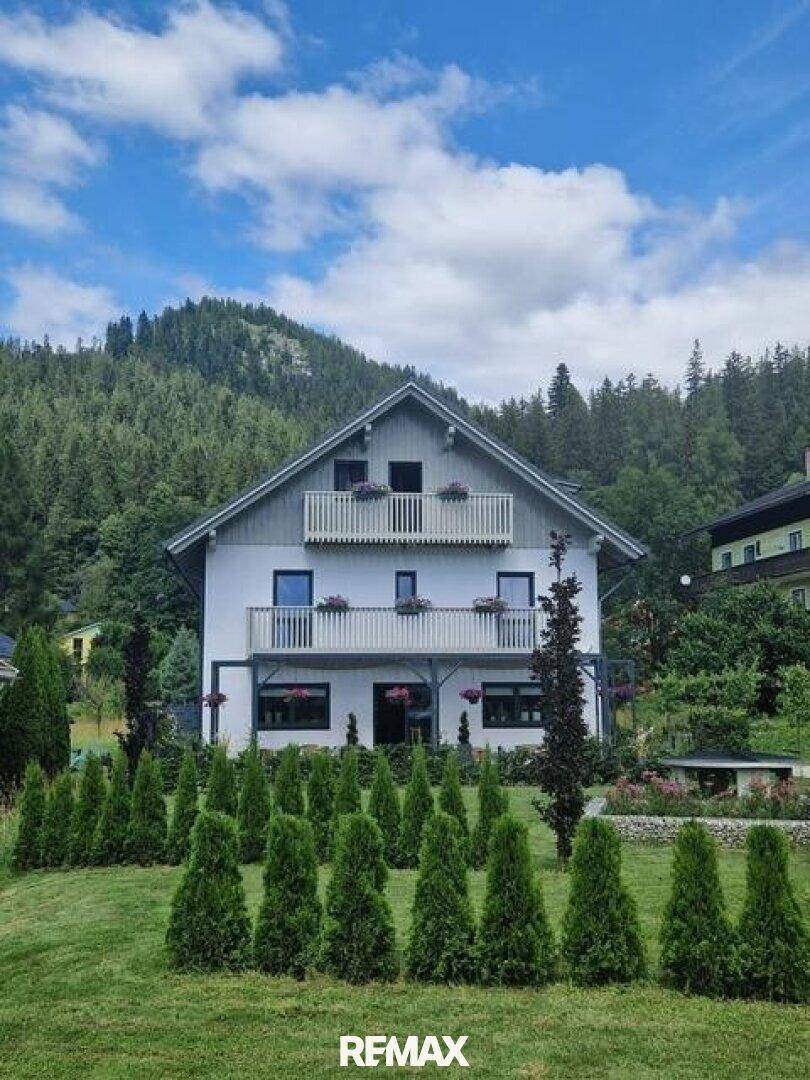 Weißes Haus mit Balkonen und einem ordentlichen Garten, eingebettet in eine bergige Landschaft.