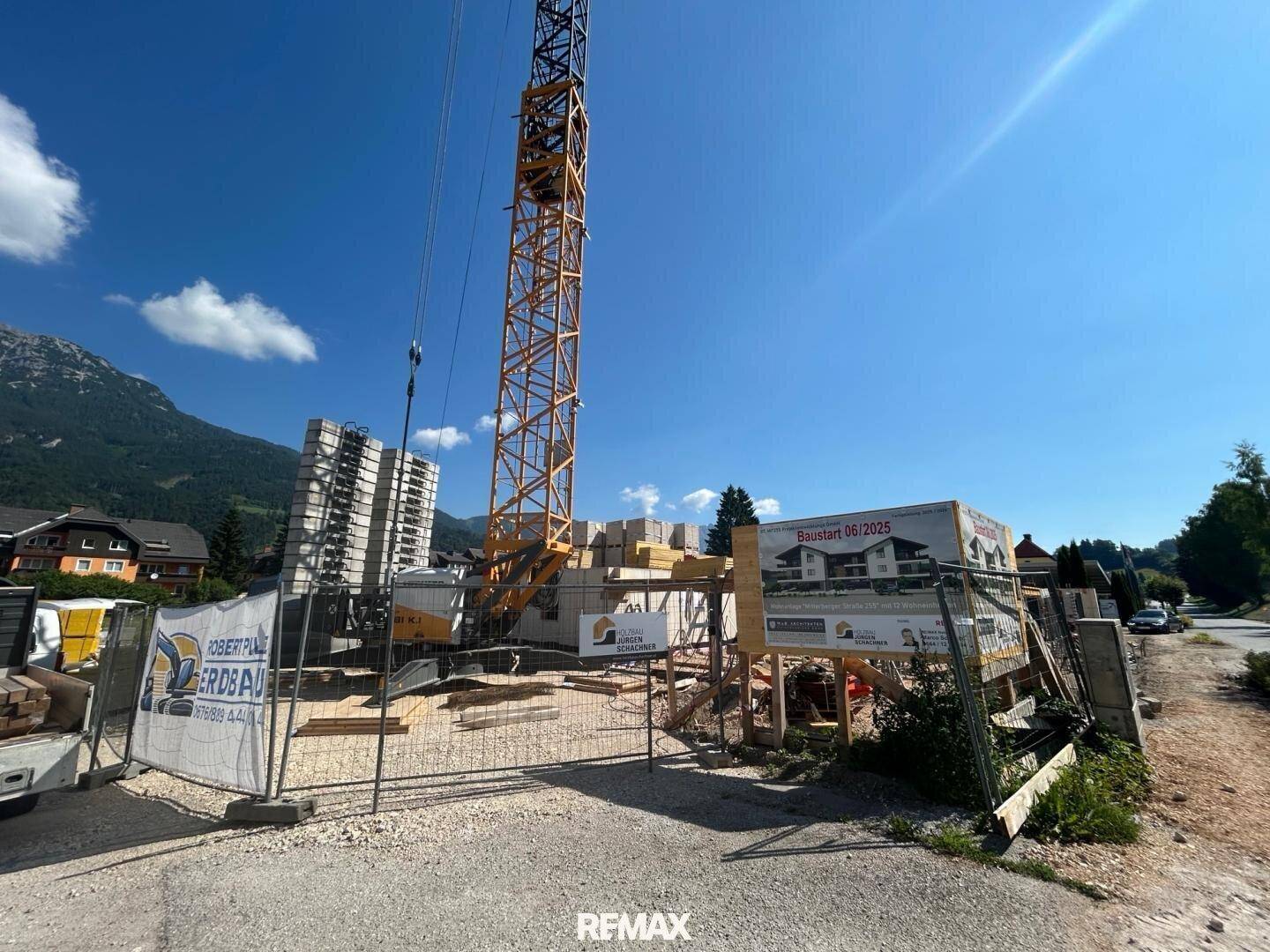 Baustelle mit Informationsschild zum Neubauprojekt und Kran unter blauem Himmel.