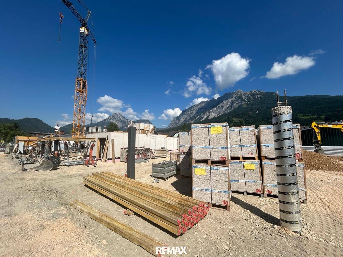 Baustelle mit Baumaterialien, Holzstapeln und Kran vor einer malerischen Berglandschaft.