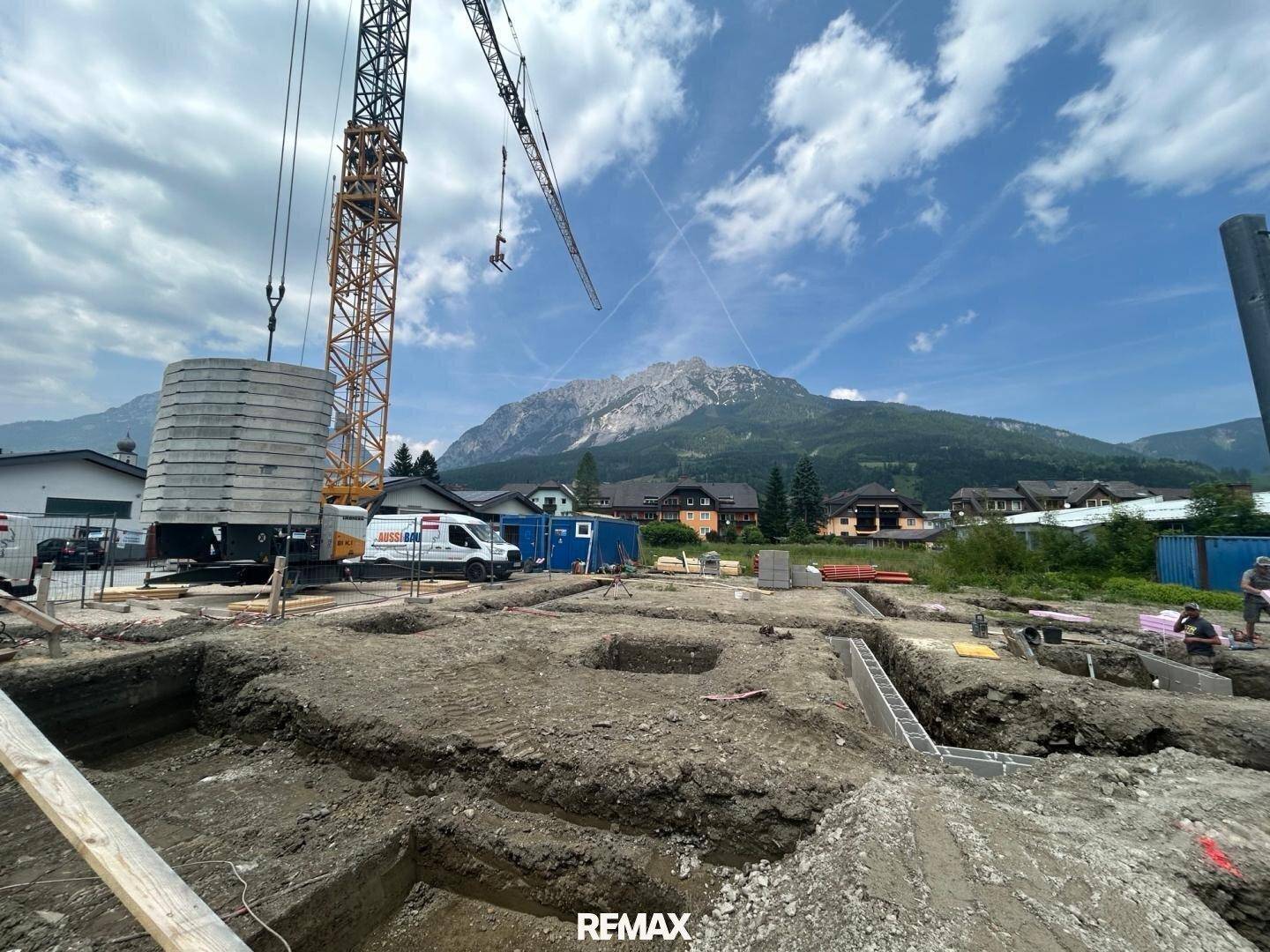 Baustelle mit ausgehobenen Fundamenten und Kran, umgeben von Bergen unter blauem Himmel.