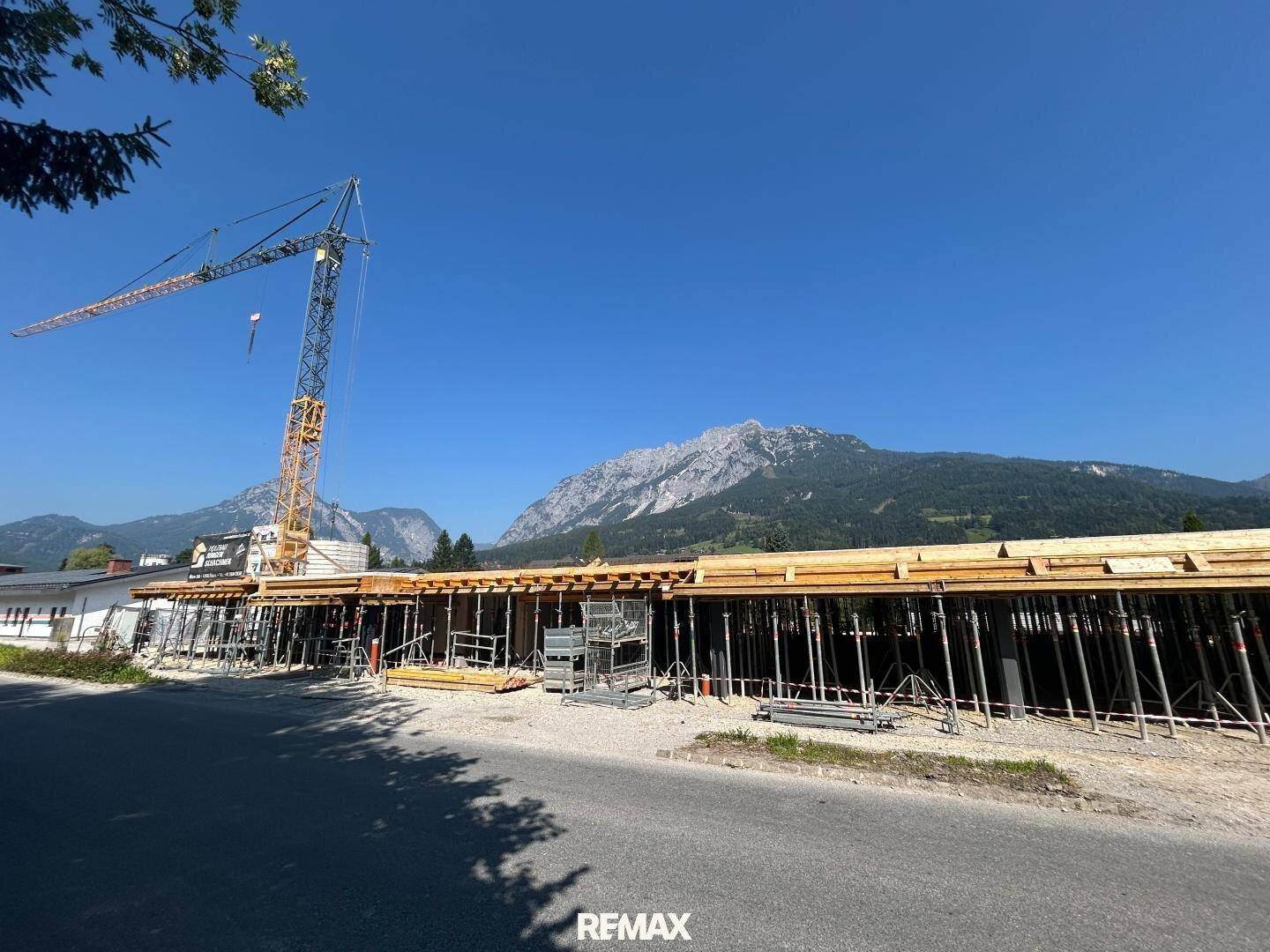 Weitläufige Baustelle mit Kran und Schalung, umgeben von grünen Bergen unter strahlendem Himmel.