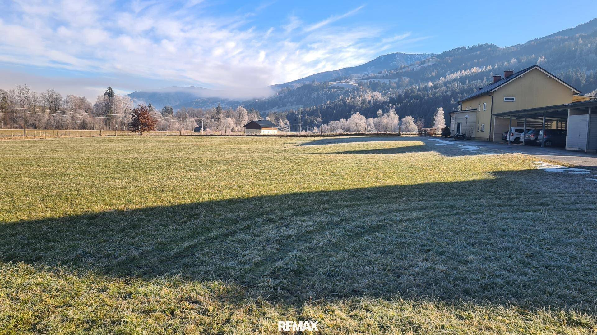 Weitläufiges grünes Feld mit einem Gebäude und Carports im Hintergrund, umgeben von frostiger Landschaft.