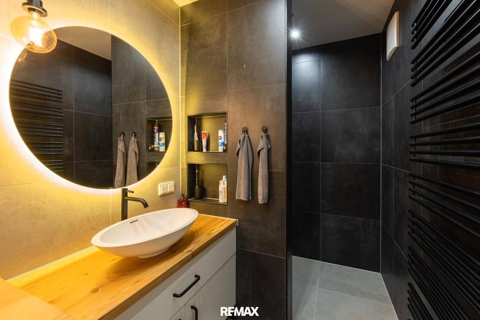 Modernes Badezimmer mit dunklen Fliesen, beleuchtetem Spiegel und Einzelwaschbecken.