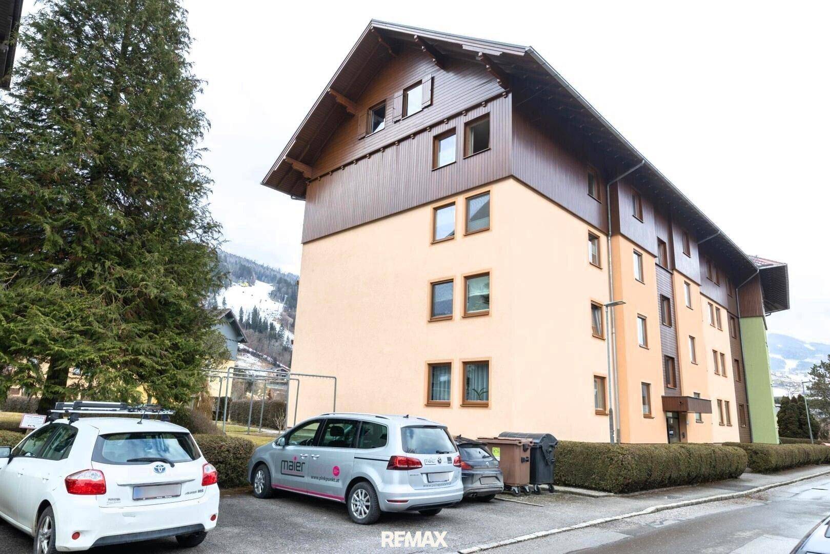 Mehrfamilienhaus mit Parkplätzen und Blick auf die Berge.