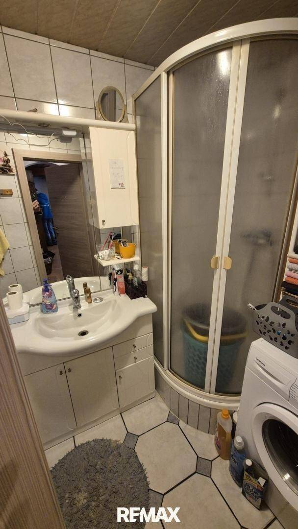 Praktisches Badezimmer mit Duschkabine, Waschbecken und Waschmaschine auf Fliesenboden.