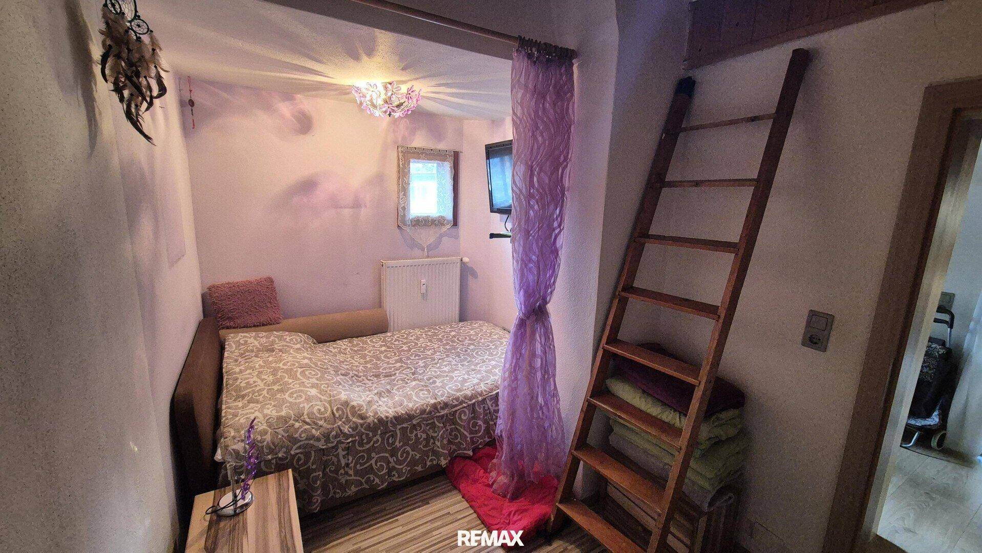 Kleines Schlafzimmer mit Bett, lila Vorhang und einer Leiter zum oberen Schlafbereich.