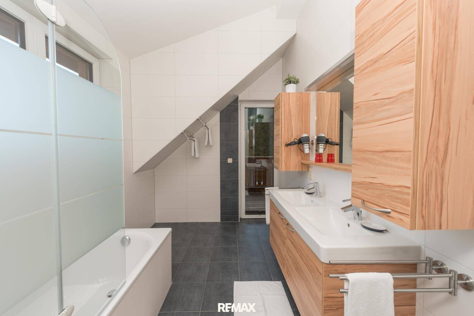 Modernes Badezimmer mit Badewanne, Doppelwaschbecken und Holzmöbeln unter der Dachschräge.