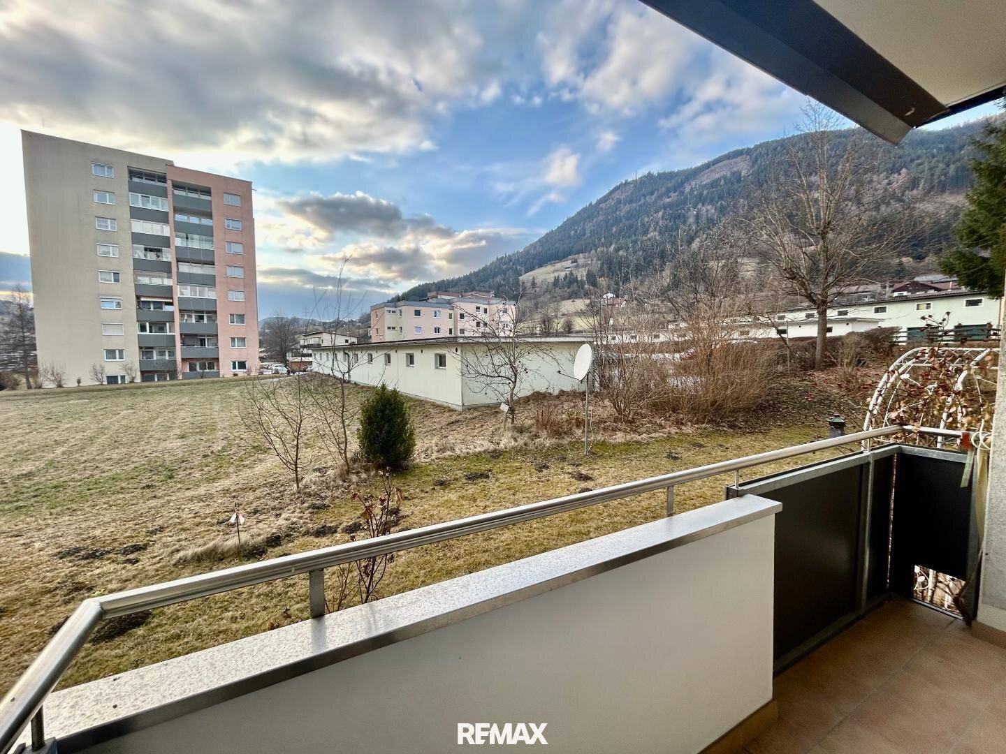 Weitläufiger Blick vom Balkon über eine Wiese auf die umliegende Berglandschaft und ein Wohnhaus.