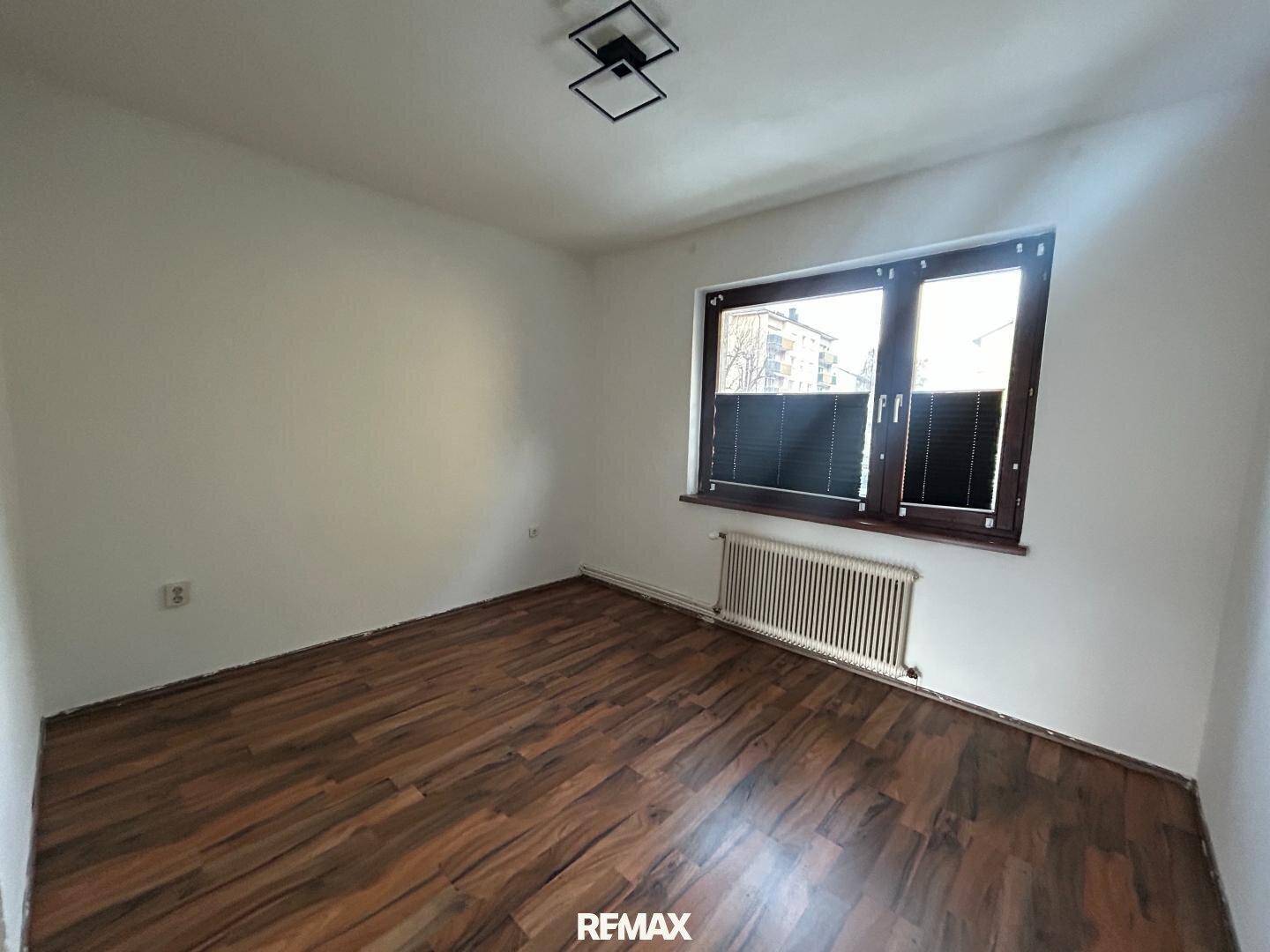 Leeres Zimmer mit dunklem Laminatboden, weißen Wänden und einem Fenster mit Jalousien.