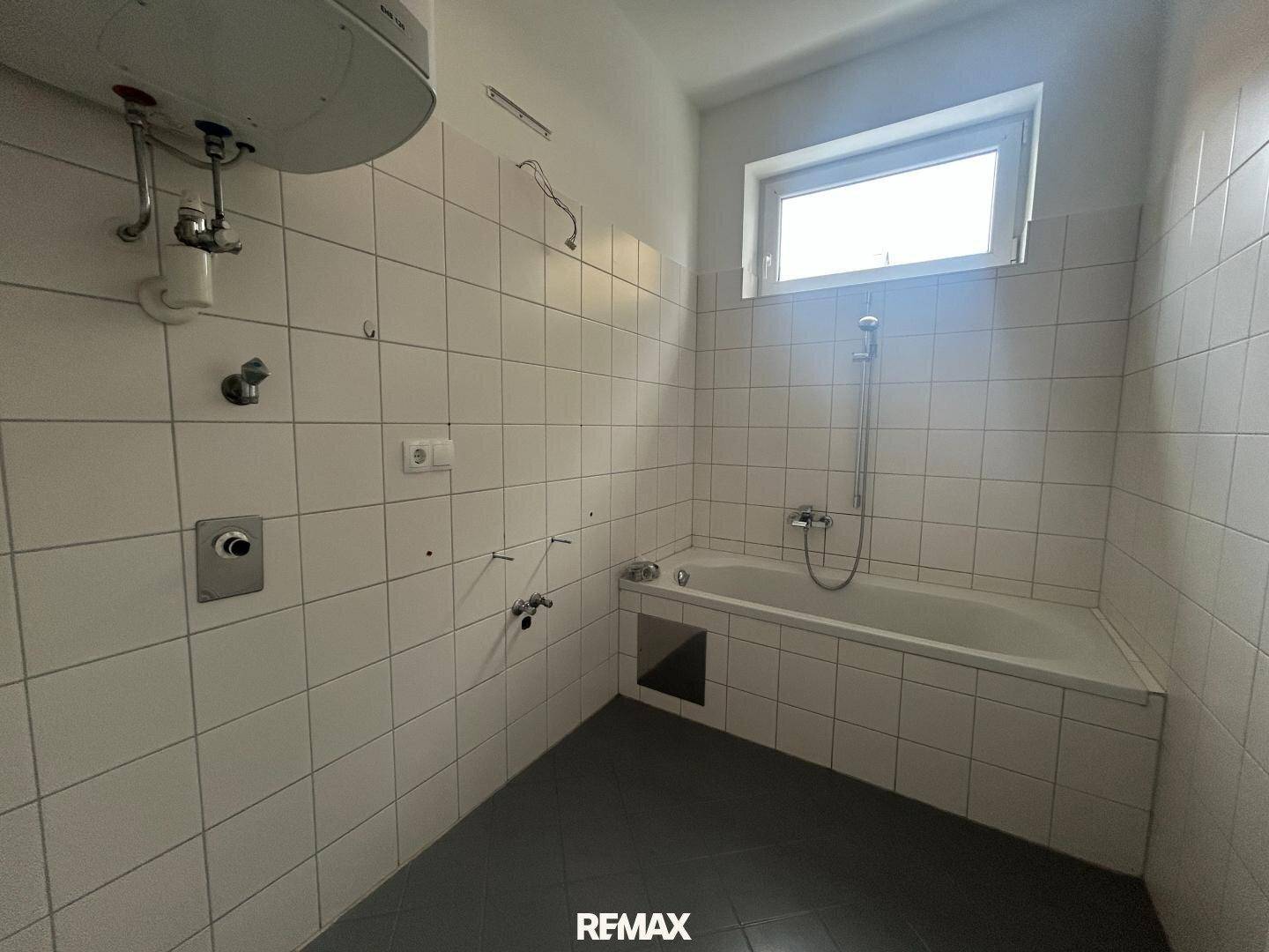 Unfertiges Badezimmer mit Badewanne, weißen Fliesen und Boiler an der Wand.
