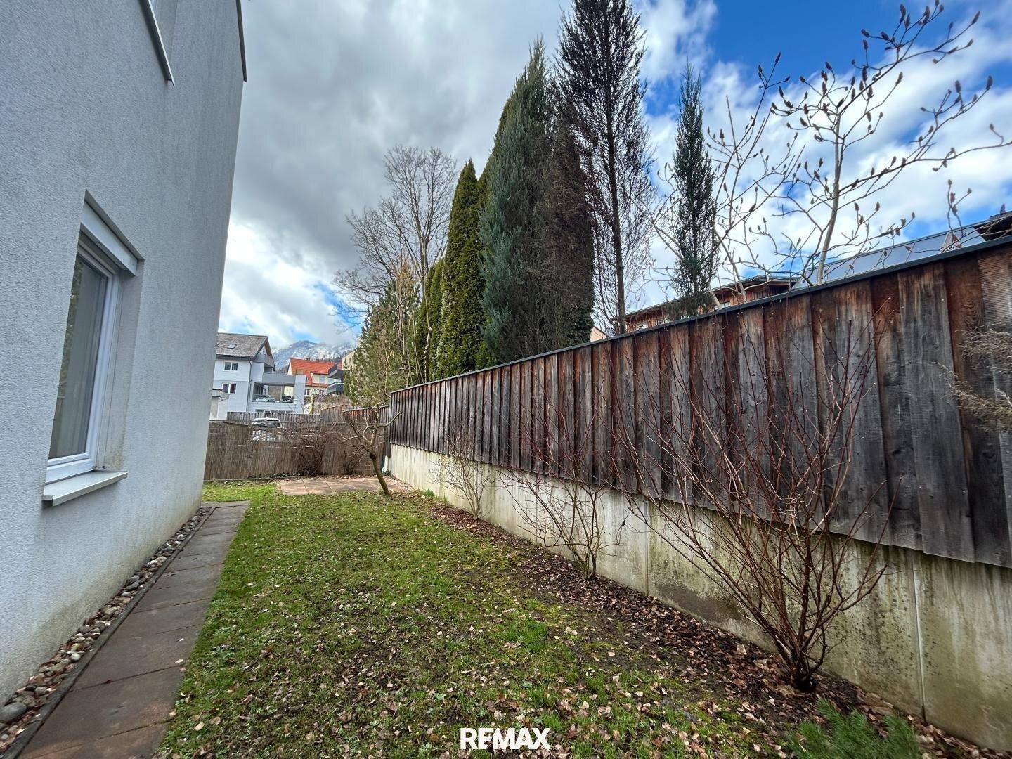 Schmaler Gartenstreifen entlang des Gebäudes mit Bäumen und Zaun.