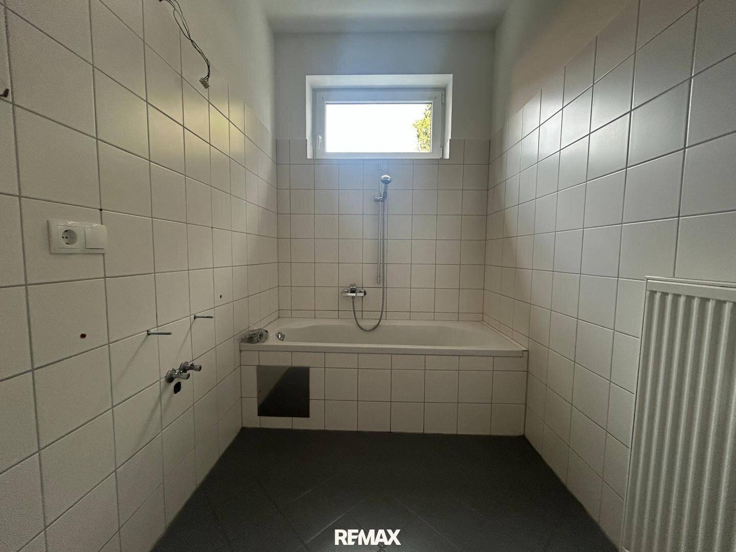 Einfaches Badezimmer mit Badewanne, weißen Wandfliesen und Fenster für Tageslicht.