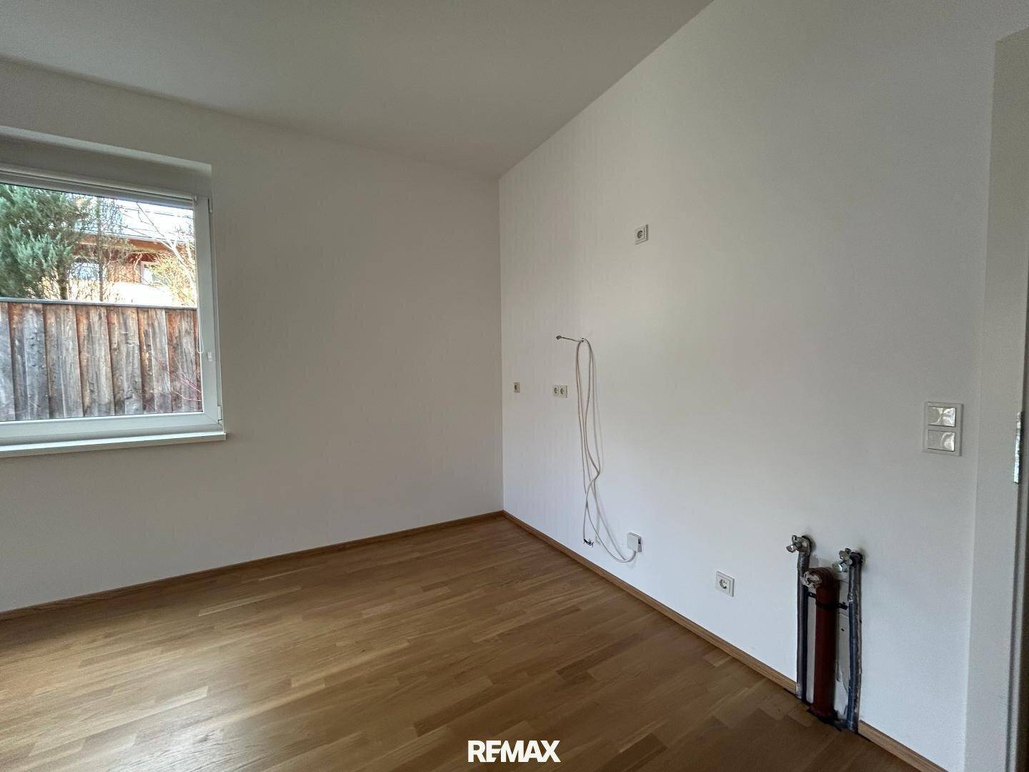 Leerer Küchenbereich mit Holzboden, Fenster und Anschlüssen für eine Einbauküche.