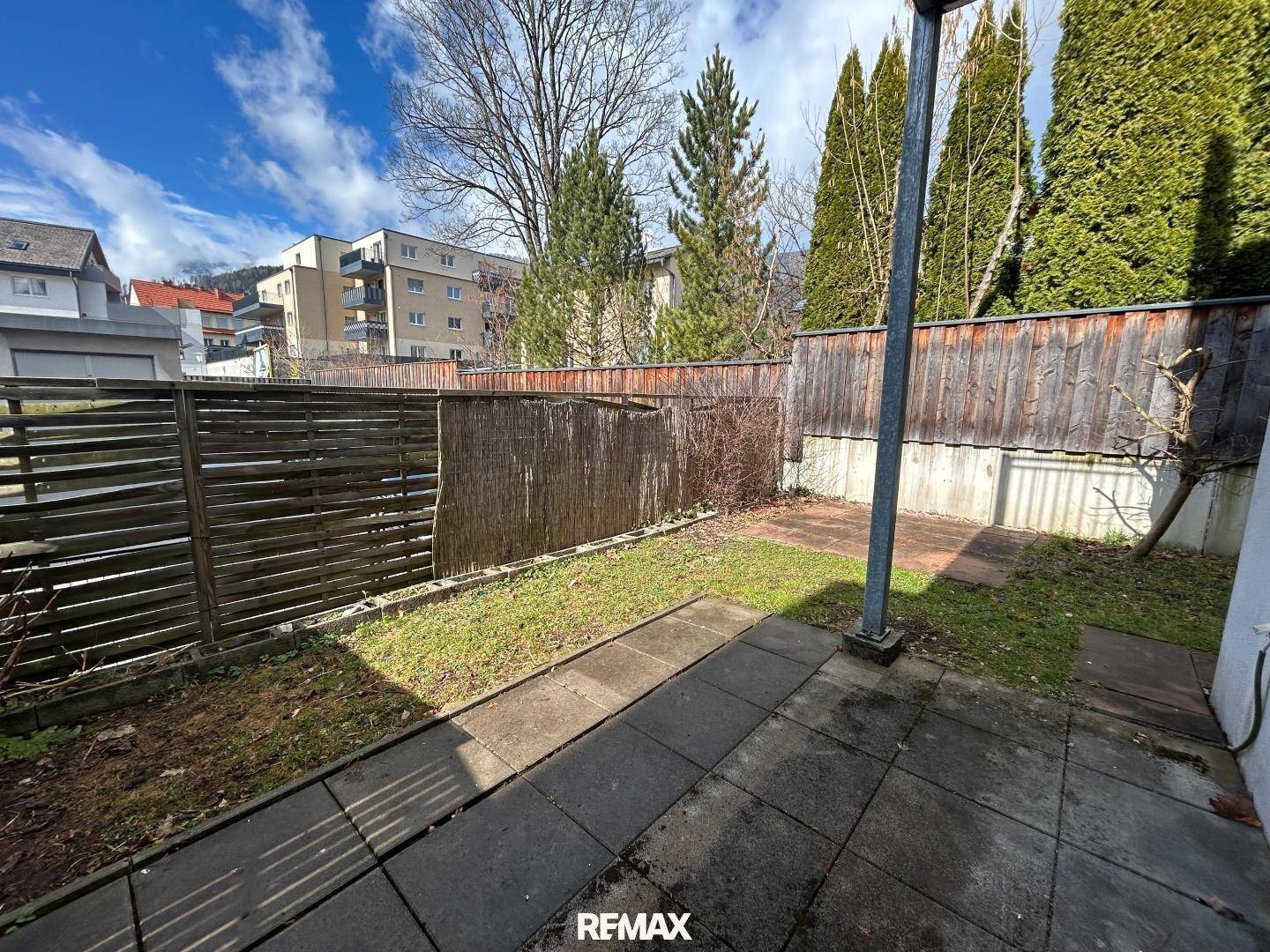 Garten mit Rasenfläche und gepflastertem Weg, umgeben von Bäumen und Zaun.