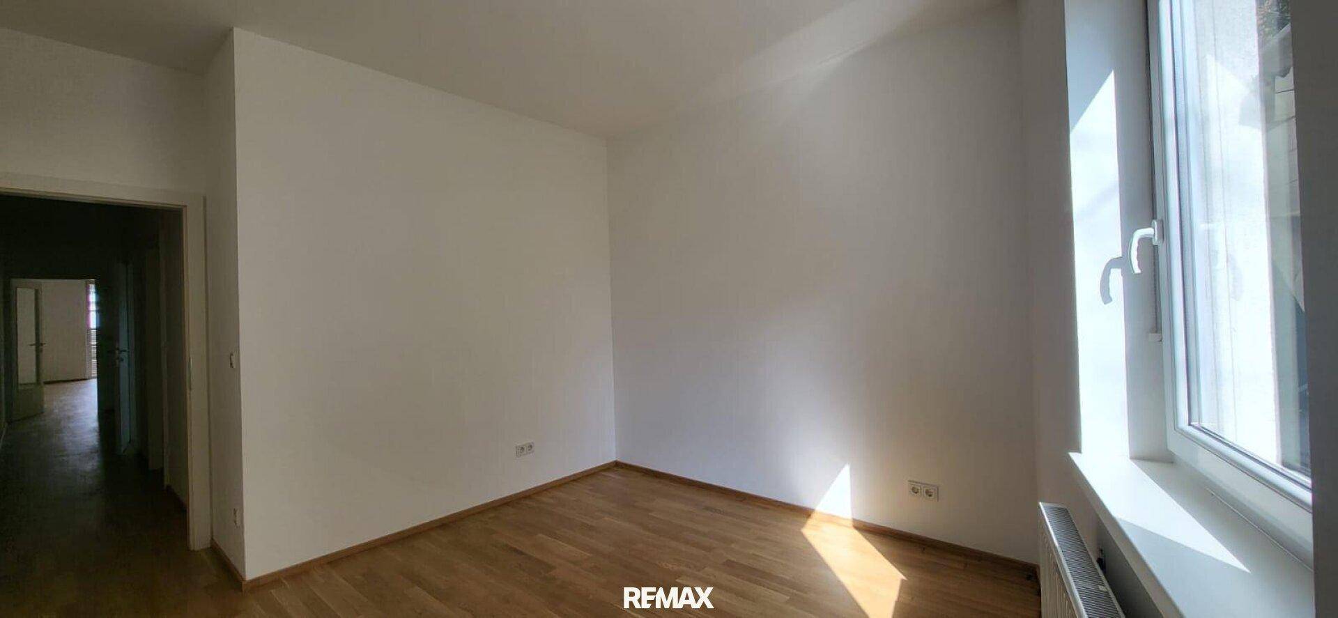 Sonniges Zimmer mit Holzboden und weißen Wänden, ein Durchgang führt zu einem weiteren Raum.