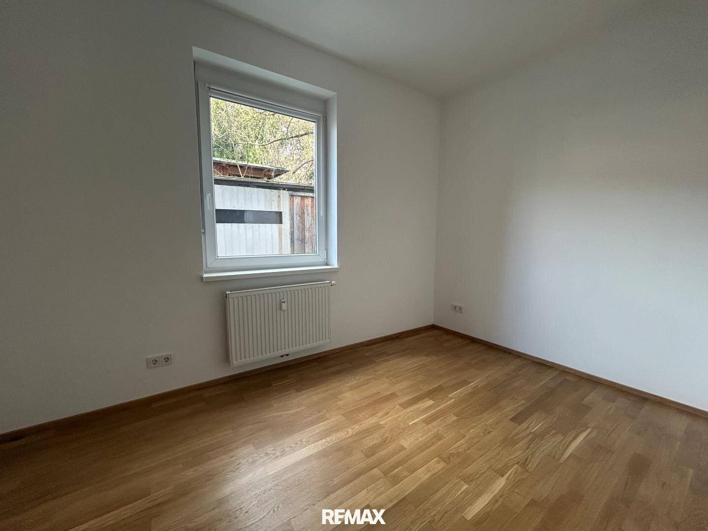 Leeres Zimmer mit hellem Holzboden, weißen Wänden und einem Fenster mit Heizkörper darunter.