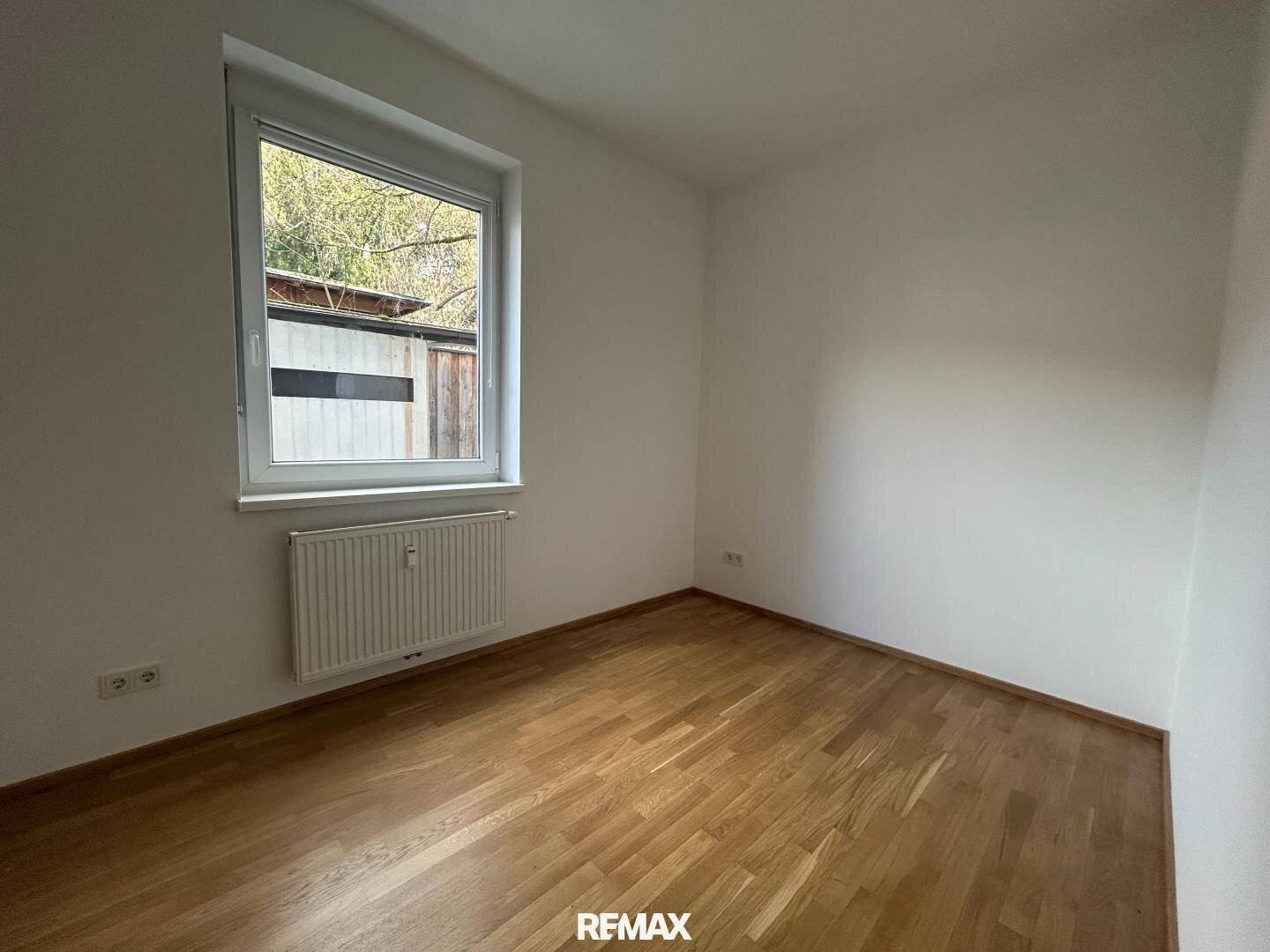 Leeres Zimmer mit hellem Holzboden, weißen Wänden und einem Fenster mit Heizkörper darunter.