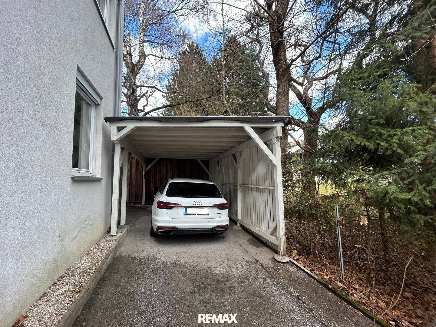 Carport mit weißem Auto und Holzwänden, umgeben von Bäumen.