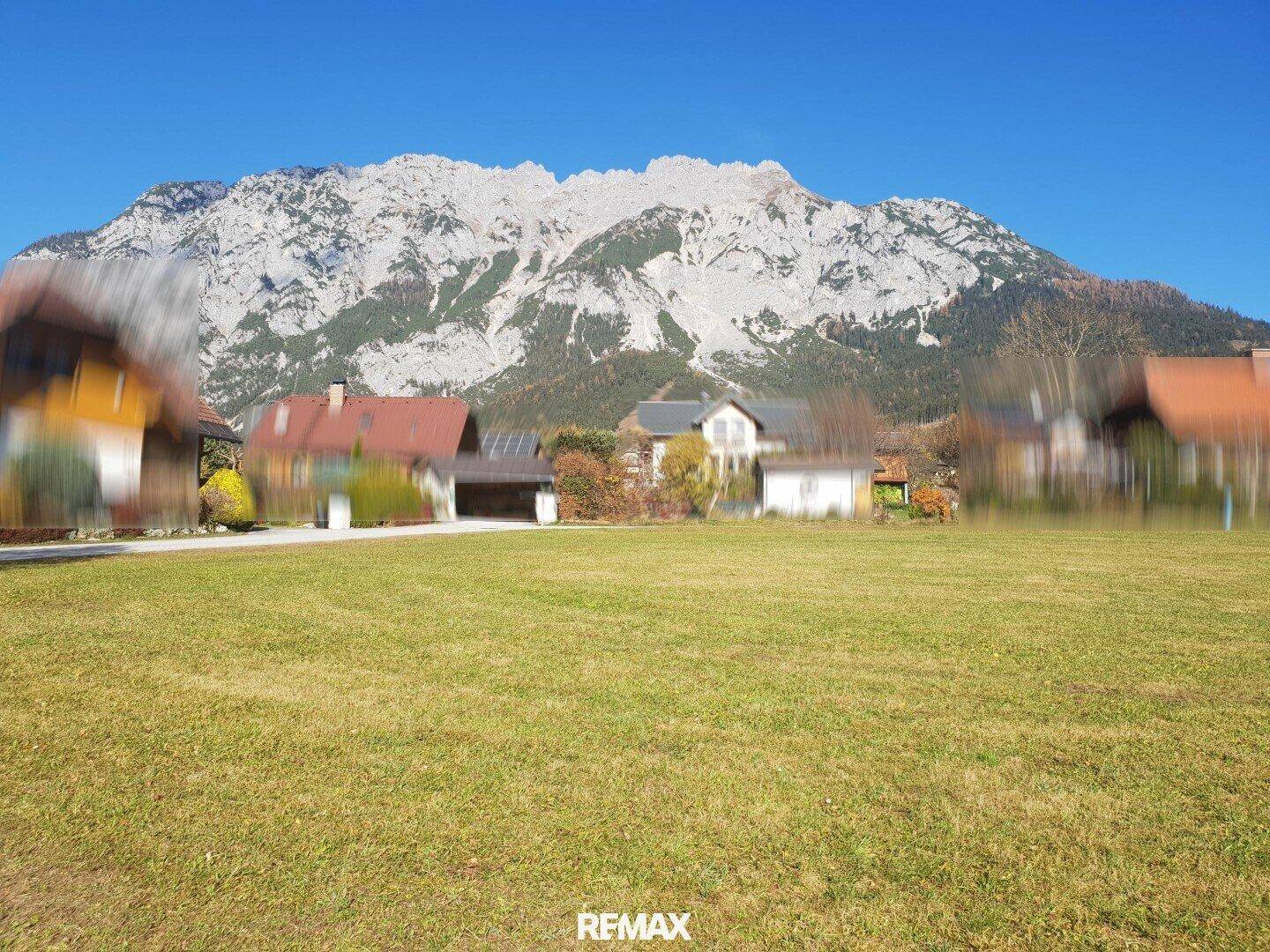 Sonnige Wiesenlandschaft mit Häusern und einem majestätischen Berg im Hintergrund unter blauem Himmel.