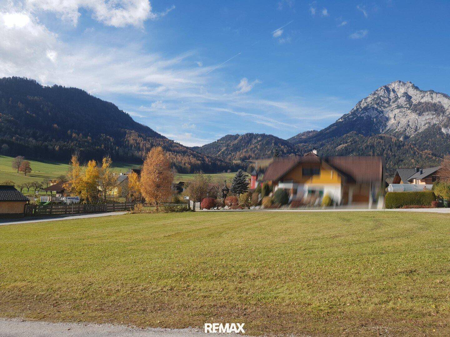 Weitläufige grüne Wiese mit Häusern und Bergen im Hintergrund unter einem bewölkten Himmel.