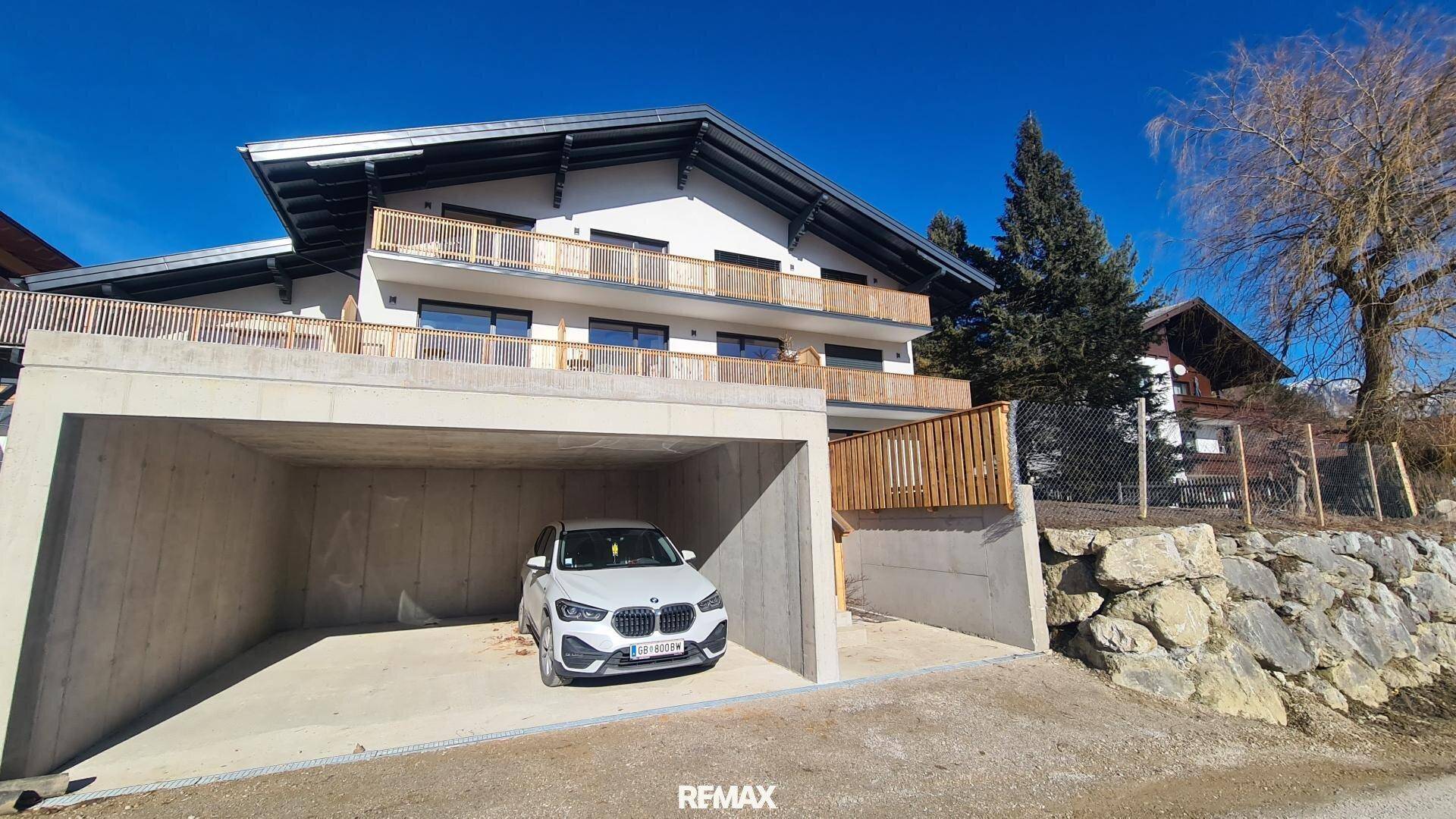 Neubau mit Carport und Balkonen in alpiner Landschaft unter strahlend blauem Himmel.