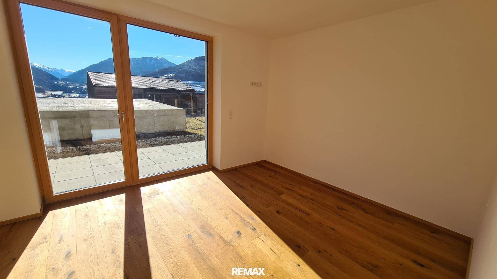 Heller Wohnraum mit Holzboden und großen Fenstern, die direkten Zugang zur Terrasse und Bergblick bieten.