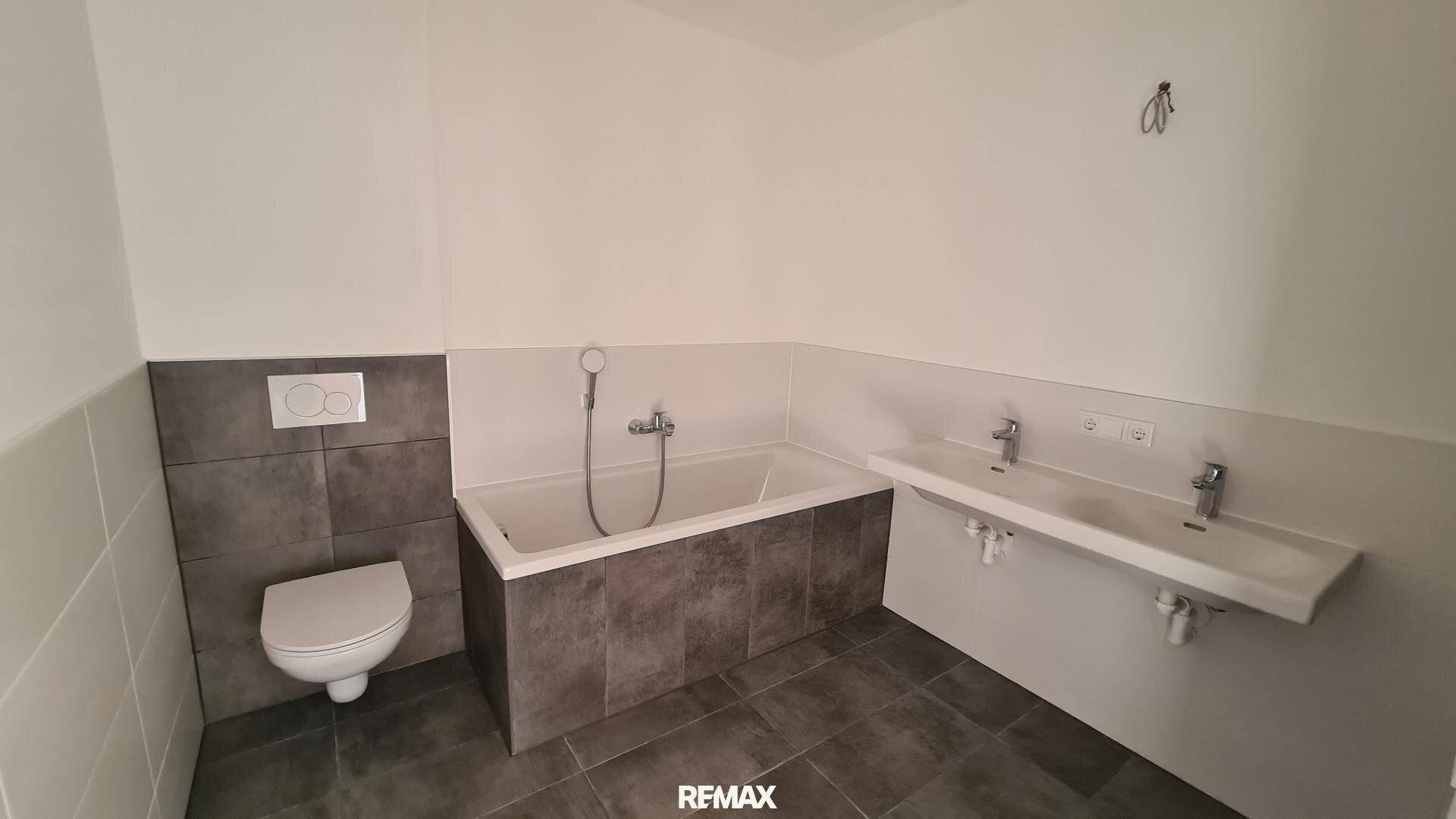 Modernes Badezimmer mit Badewanne, Toilette, Doppelwaschbecken und grauen Fliesen.