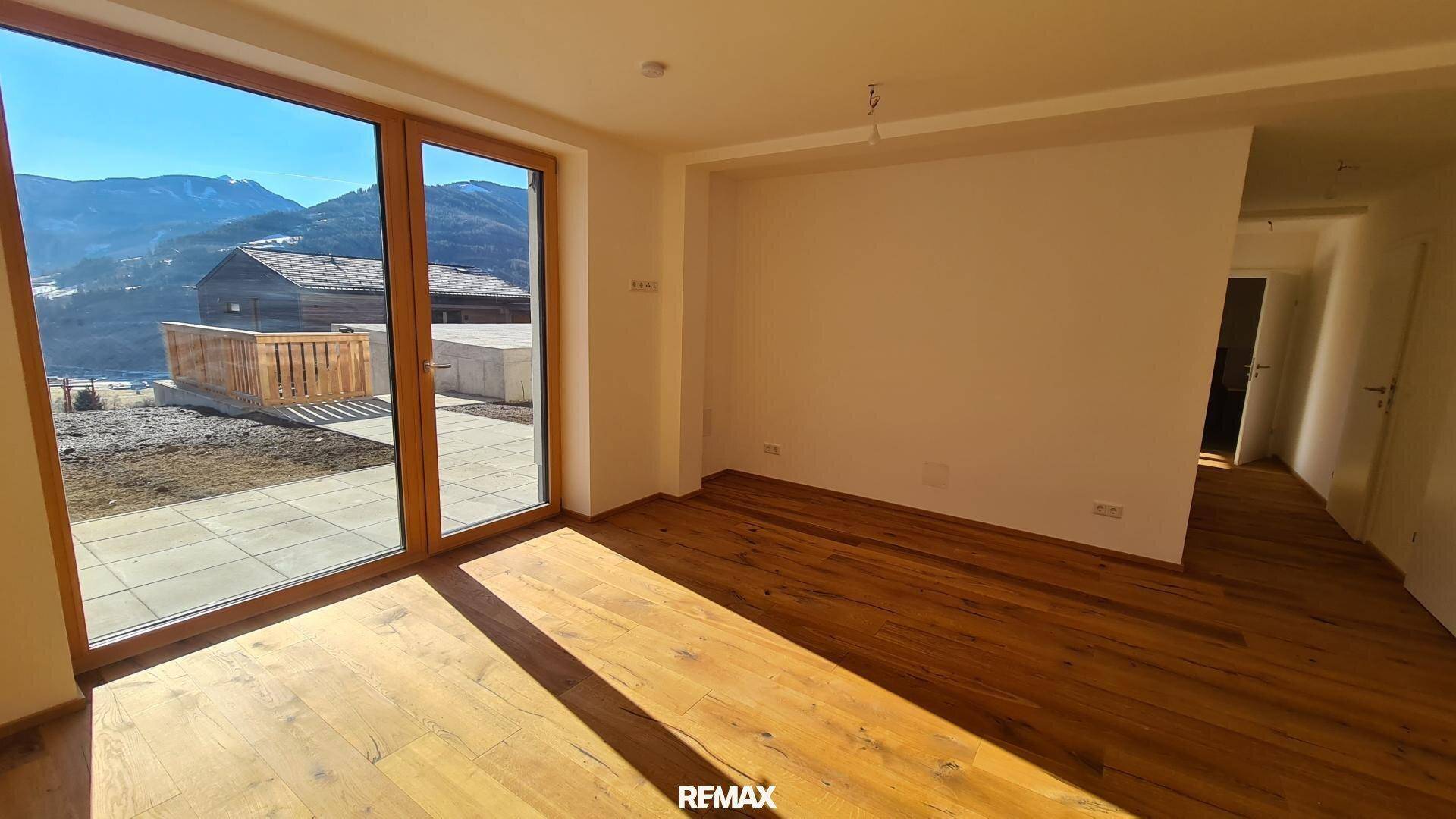 Lichtdurchfluteter Raum mit Holzboden und großen Fenstern, die Zugang zur Terrasse und Bergblick bieten.