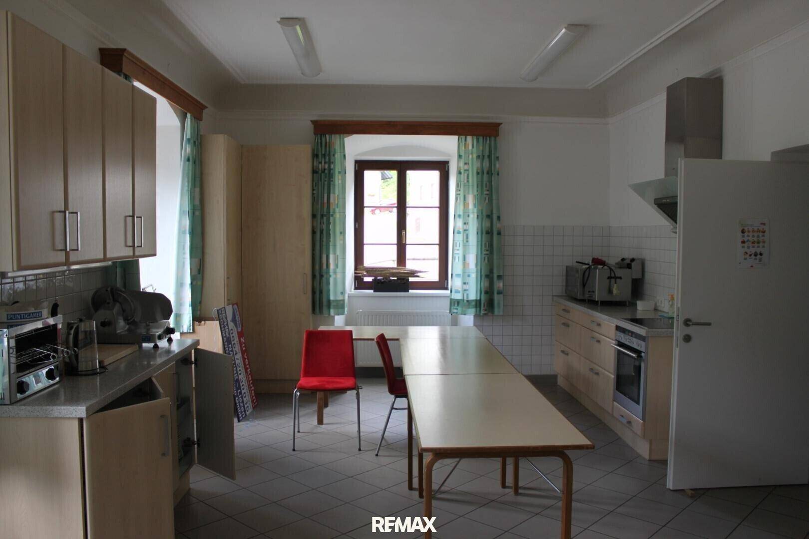 Funktionale Küche mit hellen Holzschränken, Fliesenboden und einem Fenster mit Vorhängen.