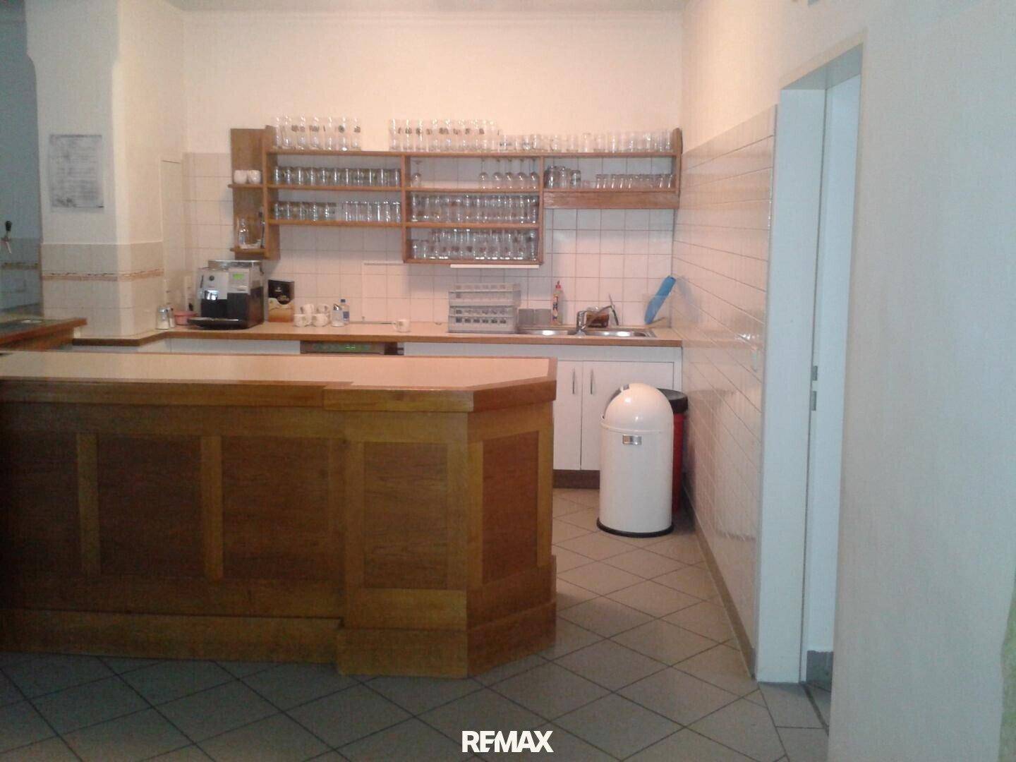 Bar-Bereich mit Holztheke, Fliesenboden und Regalen für Gläser, in einem gut erhaltenen Zustand.