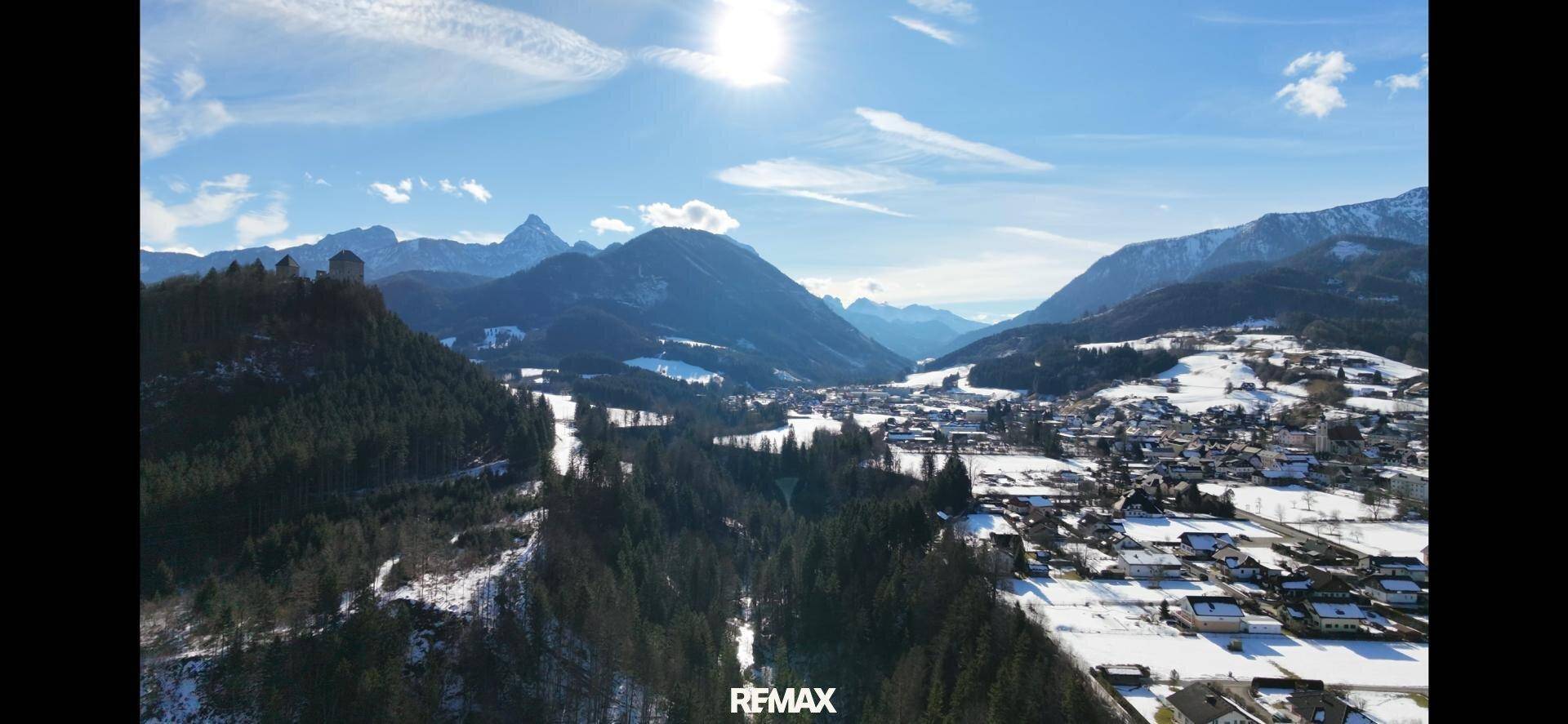 Weitläufige Luftaufnahme eines Tals mit verschneiten Bergen, Wald und einem Dorf unter blauem Himmel.