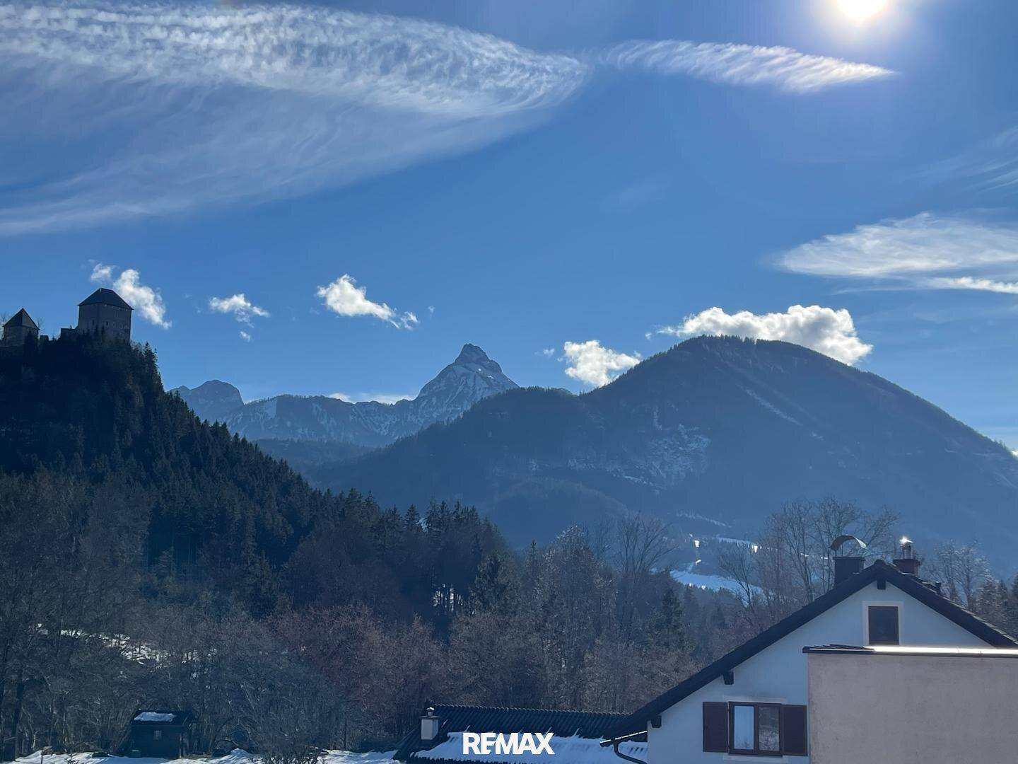Sonniger Blick auf die Berge mit einer Burg und einem Haus im Vordergrund unter blauem Himmel.