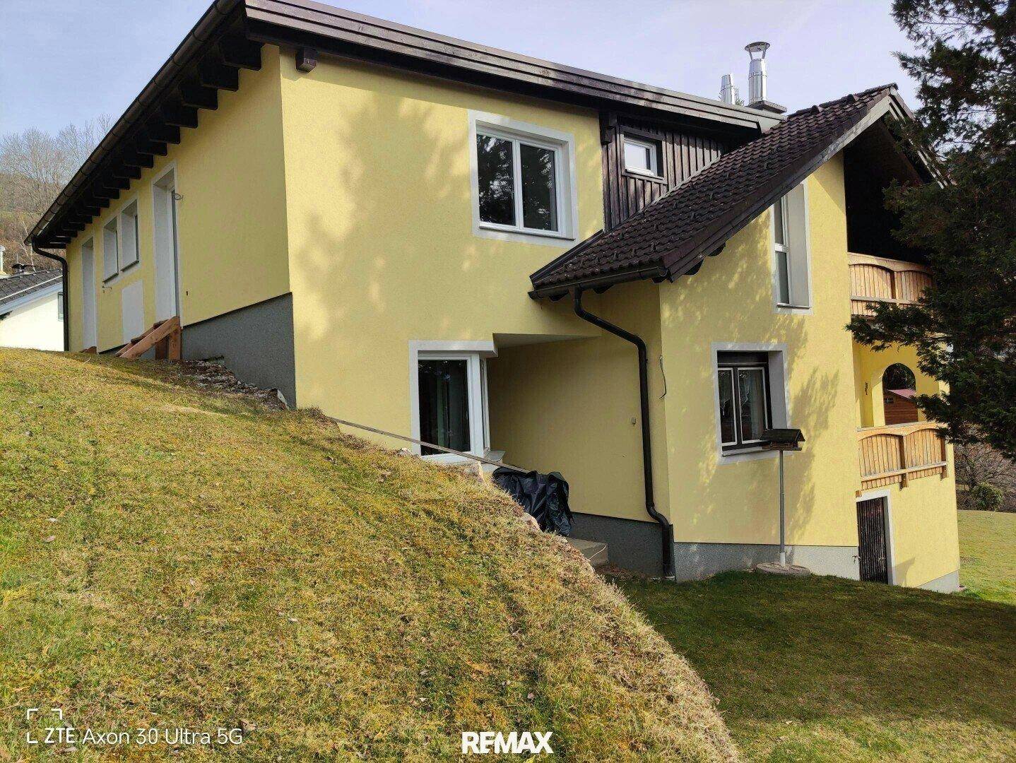 Außenansicht des Hauses mit gelber Fassade, Dach und der umliegenden Hanglage.