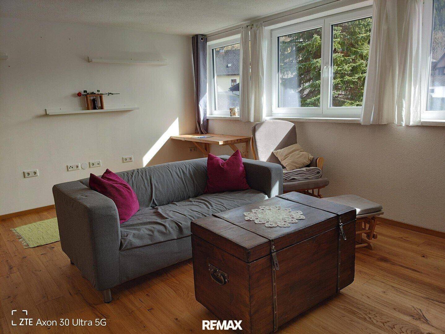 Gemütliches Wohnzimmer mit grauem Sofa, Holzboden und Fenstern mit Blick ins Grüne.