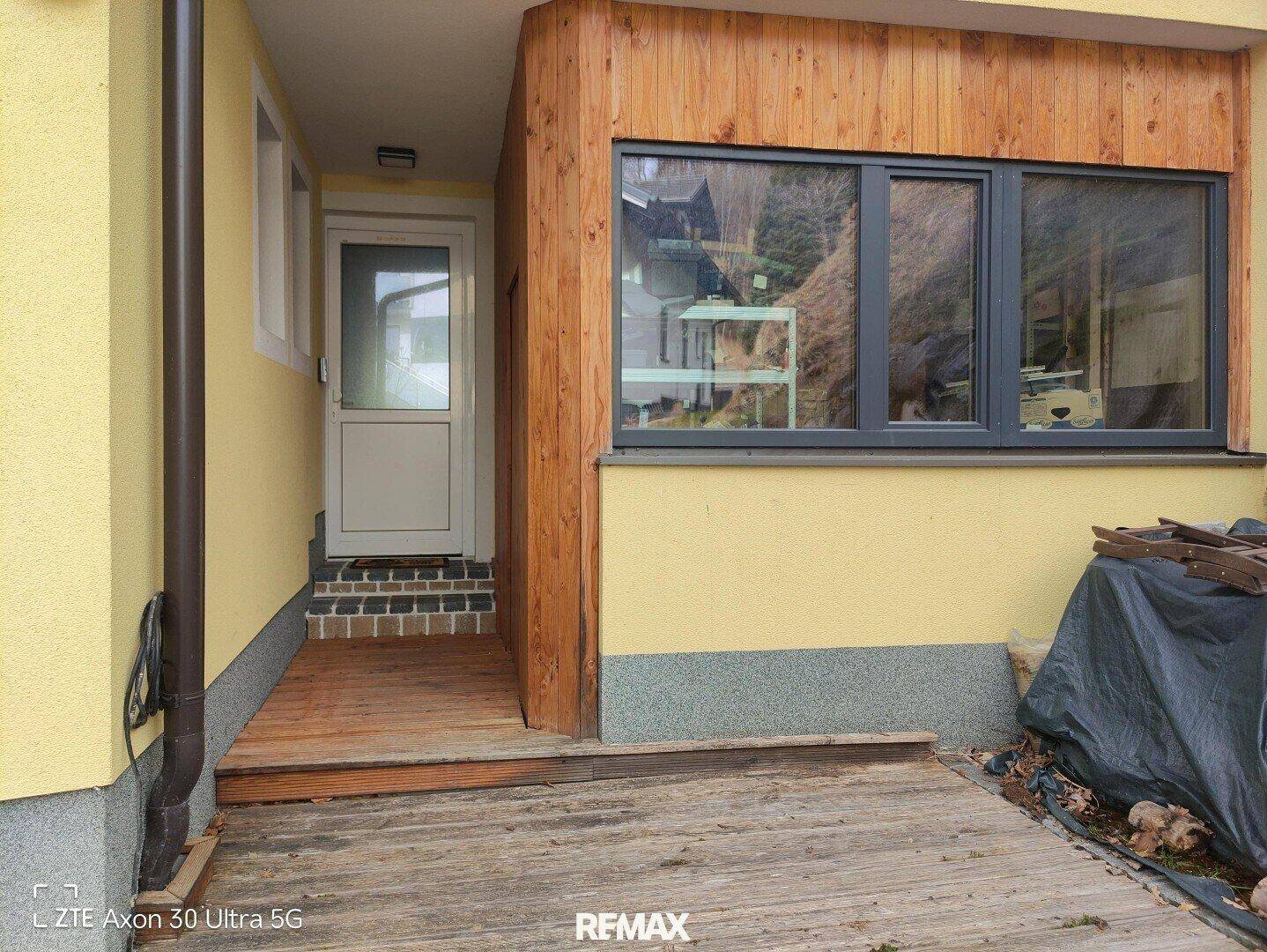 Eingangsbereich des Hauses mit gelber Fassade, Holzverkleidung und Holzterrasse.