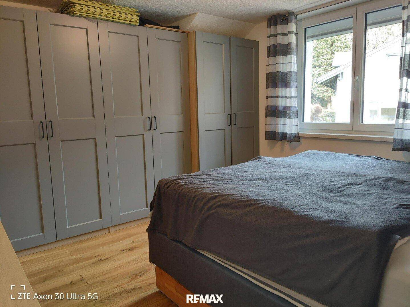 Schlafzimmer mit Bett, grauen Einbauschränken und einem Fenster mit gestreiften Vorhängen.