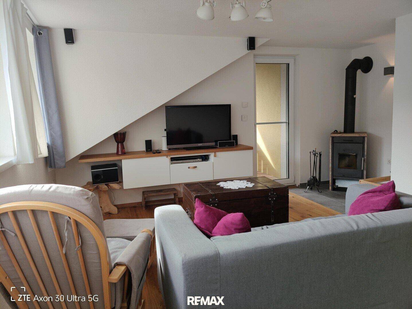 Wohnzimmer mit Kaminofen, TV-Möbeln, grauem Sofa und Zugang zu einem Außenbereich.