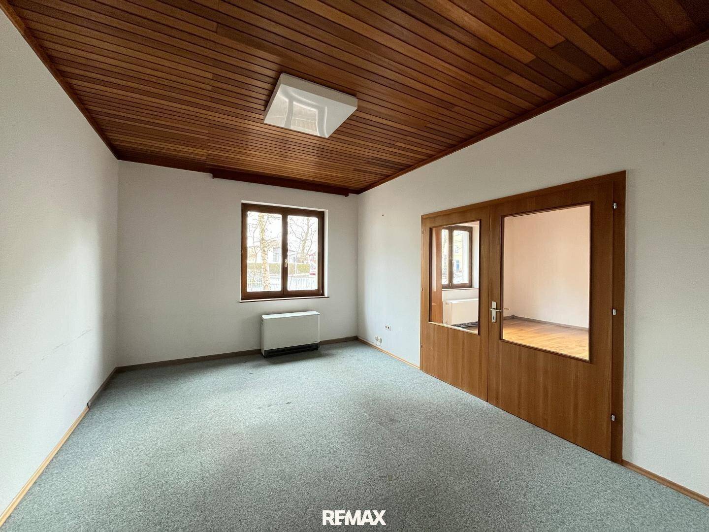 Leerer Raum mit Teppichboden, einer Holzdecke und zwei Fenstern, die Tageslicht hereinlassen.