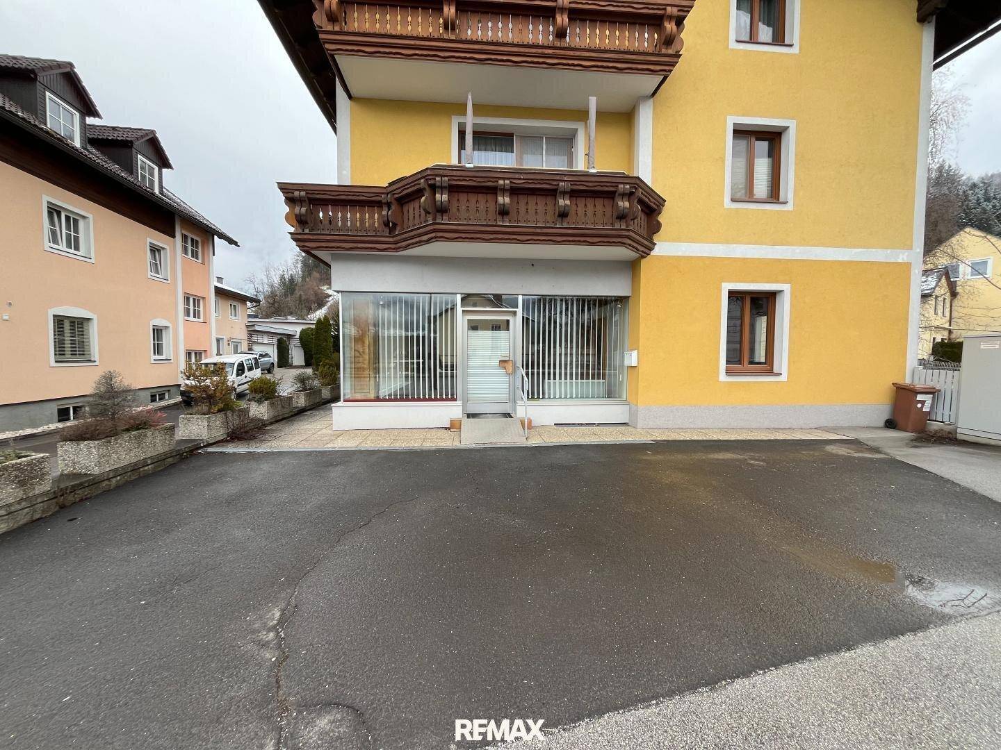 Gelbes Gebäude mit traditionellen Holzbalkonen und einer modernen Glasfassade im Erdgeschoss, umgeben von einer gepflasterten Einfahrt.