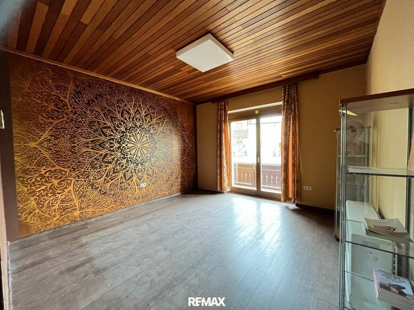 Leerer Raum mit einer auffälligen goldenen Mustertapete, Holzdecke und Zugang zu einem Balkon.