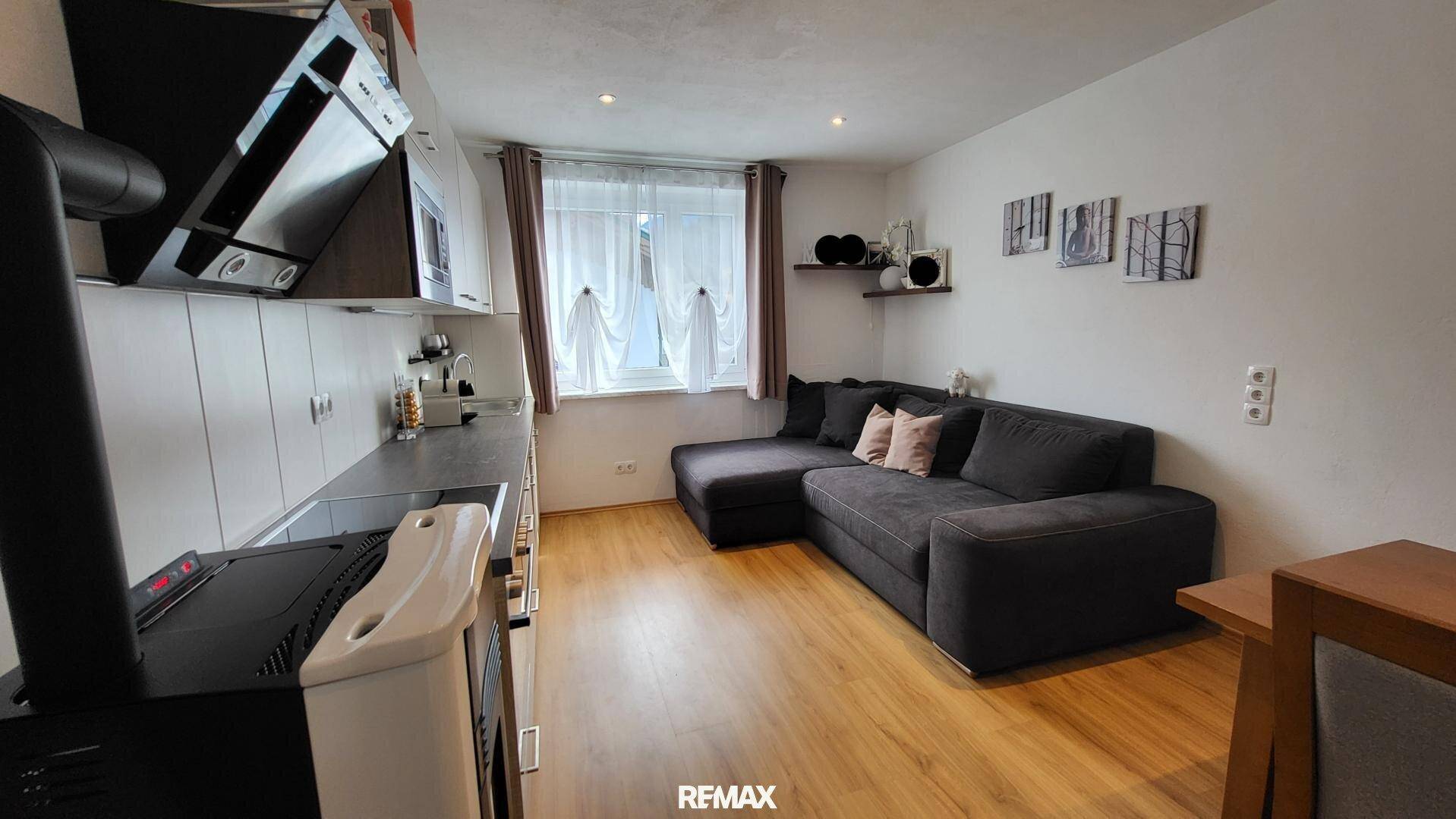Gemütlicher Wohnbereich mit L-förmigem Sofa und integrierter Küchenzeile, hell durch Fenster.
