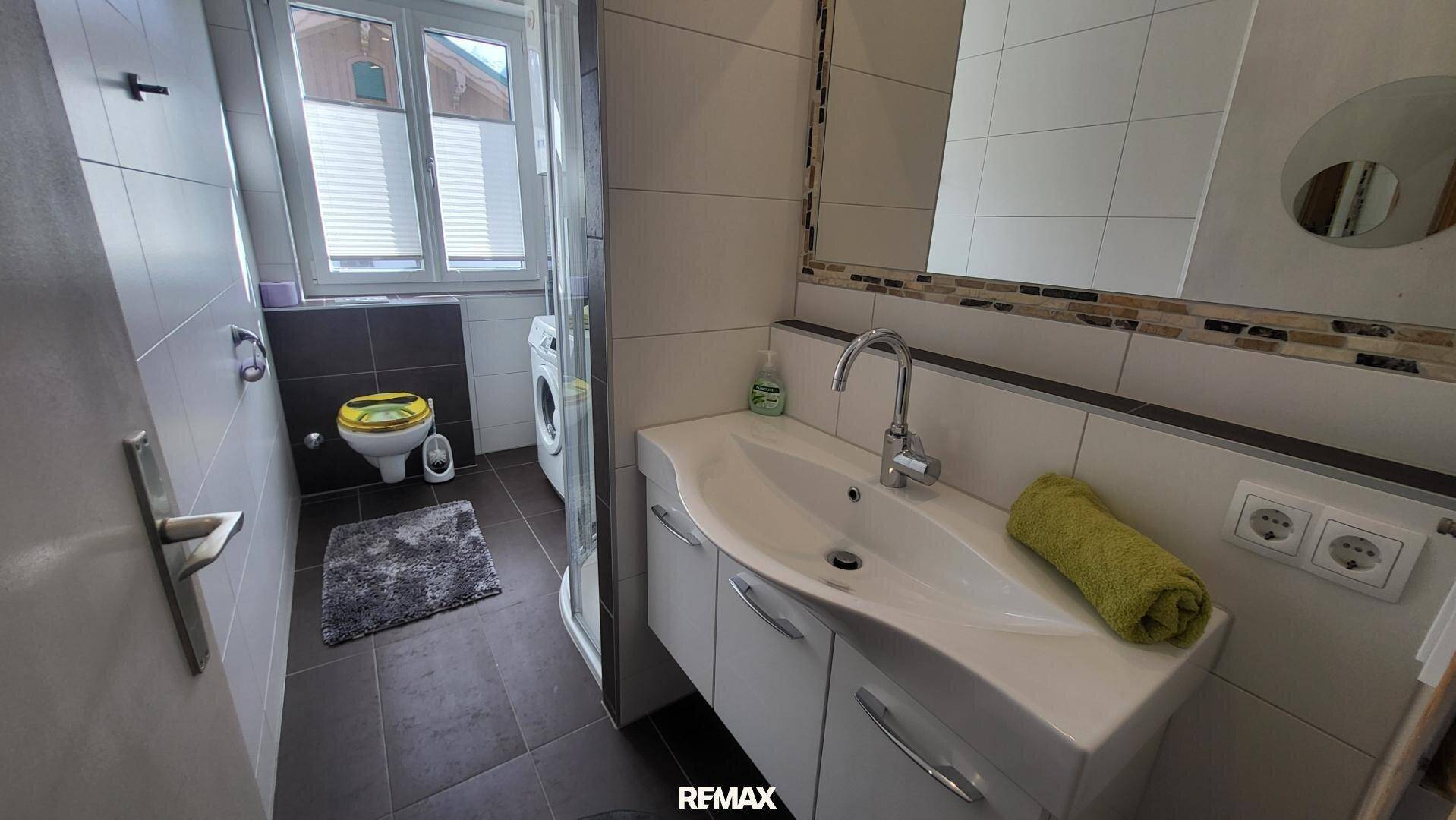Modernes Badezimmer mit Dusche, WC, Waschbecken und Waschmaschine, hell durch Fenster.
