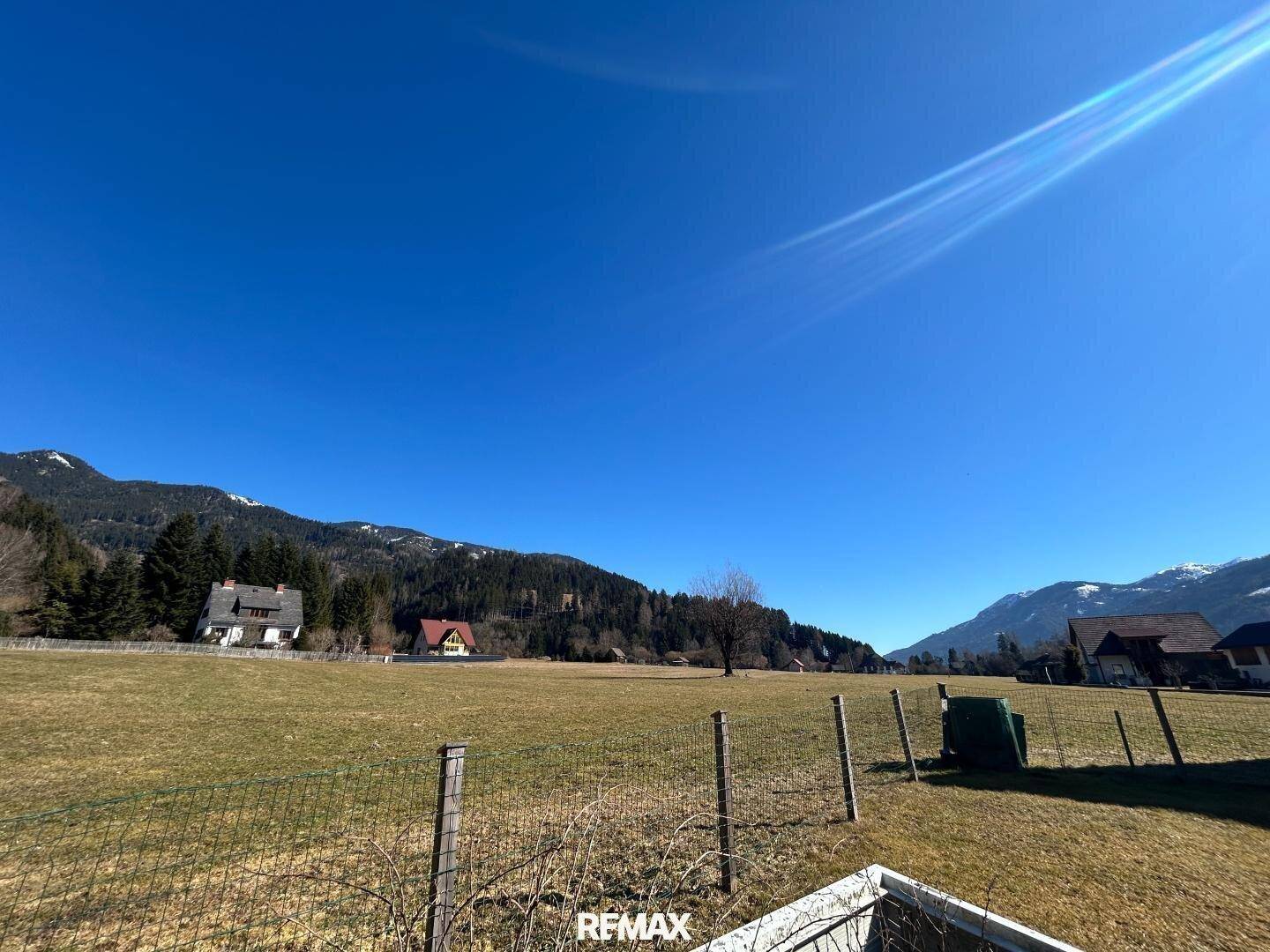 Weitläufige grüne Landschaft mit Bergen im Hintergrund und ländlicher Bebauung unter blauem Himmel.