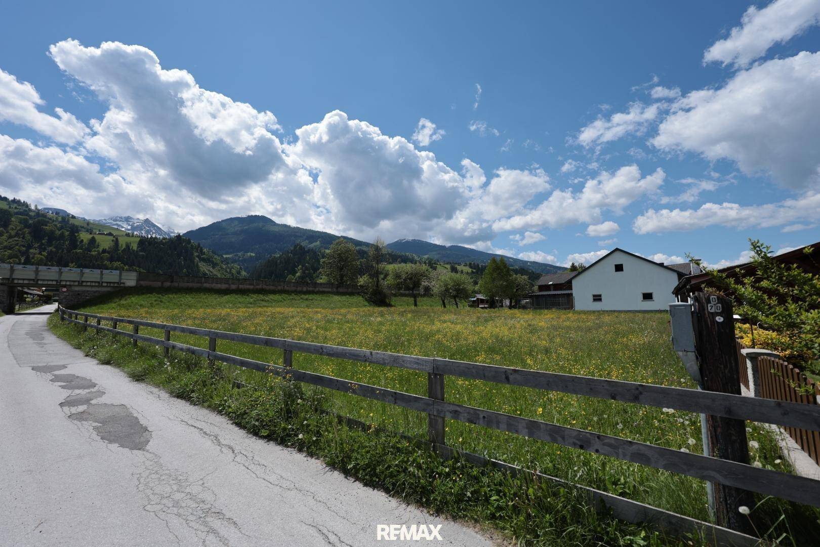 Weitläufige grüne Landschaft mit Wiesen, Bergen im Hintergrund und einem Haus in der Ferne.