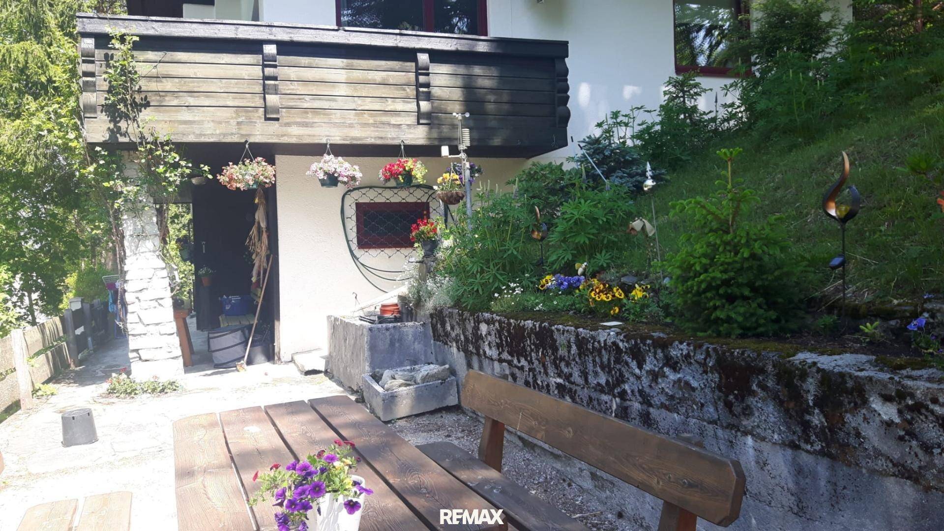 Sonniger Außenbereich mit Holztisch und Bänken, umgeben von blühenden Blumenbeeten und einem kleinen Brunnen.