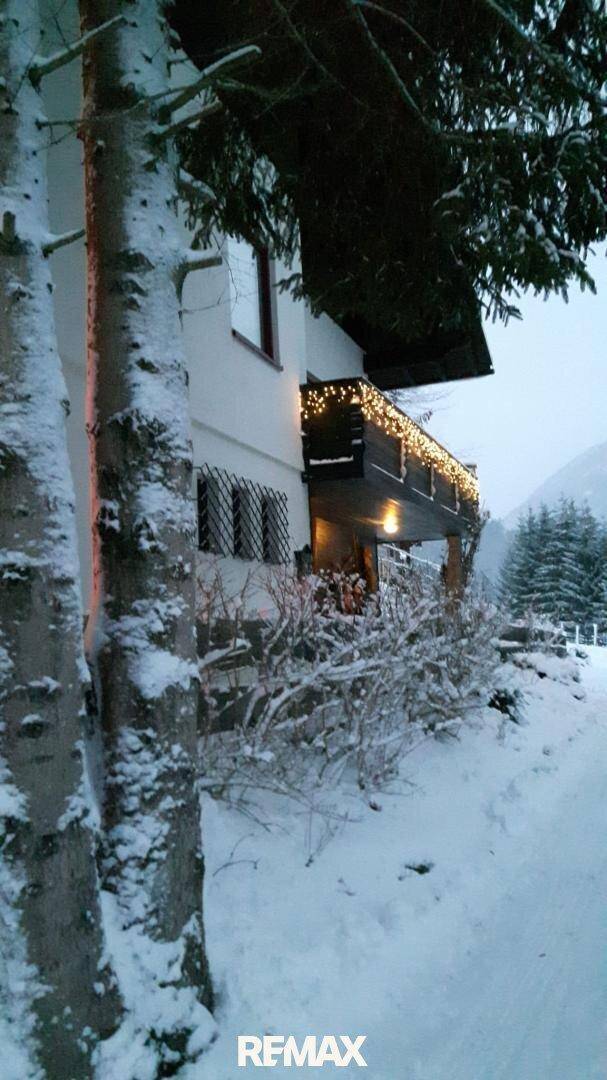 Hausfassade im Winter bei Dämmerung, geschmückt mit festlicher Lichterkette und umgeben von Schnee.