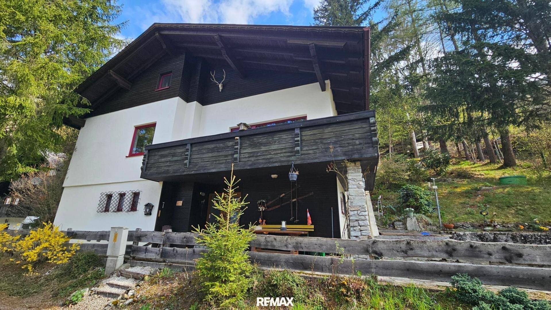Traditionelles Haus im alpinen Stil mit Holzbalkon und Hirschgeweih-Dekoration, umgeben von Bäumen.