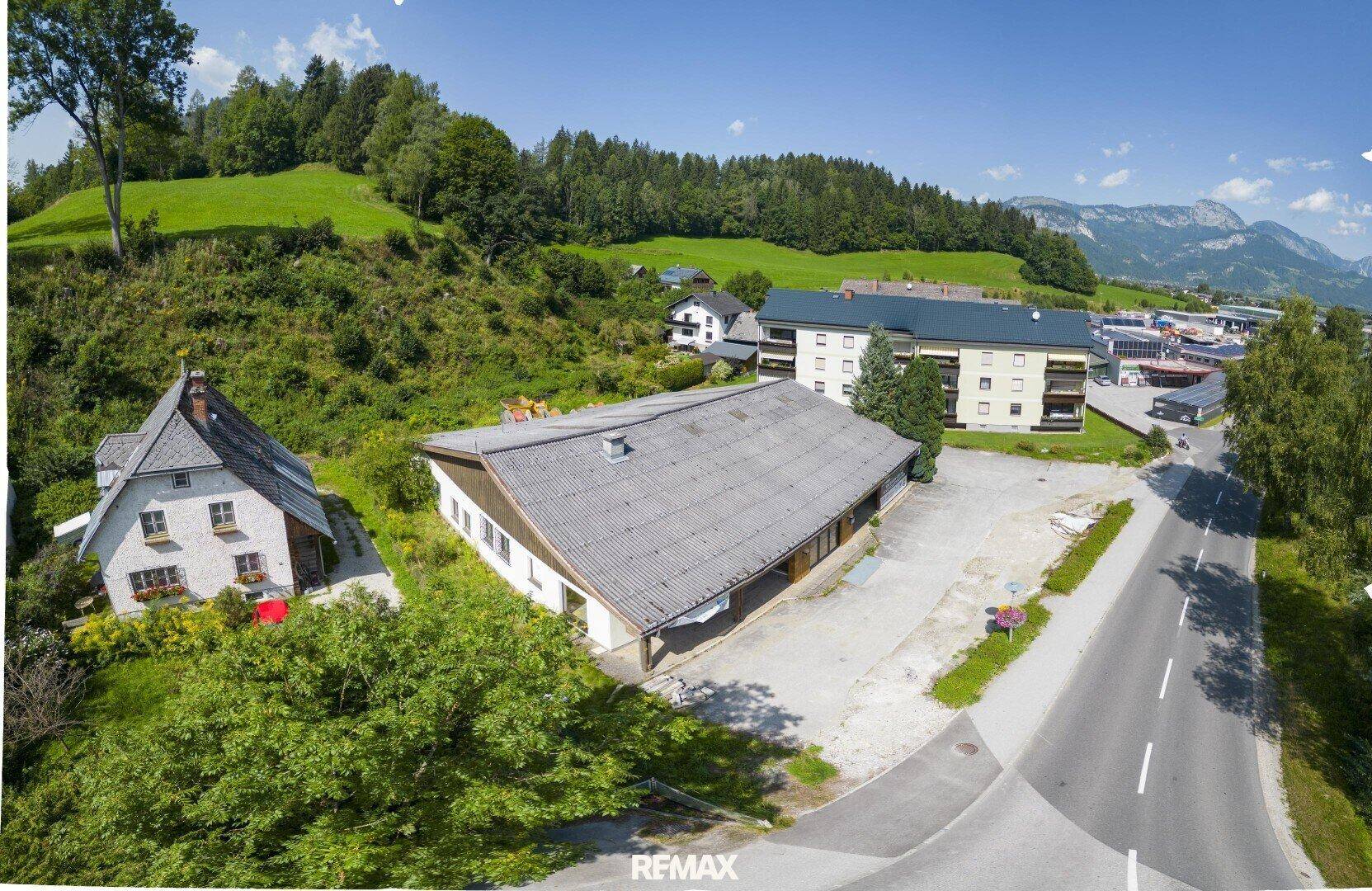 Luftaufnahme eines Gebäudes mit Satteldach und umliegender grüner Landschaft sowie Bergen im Hintergrund.